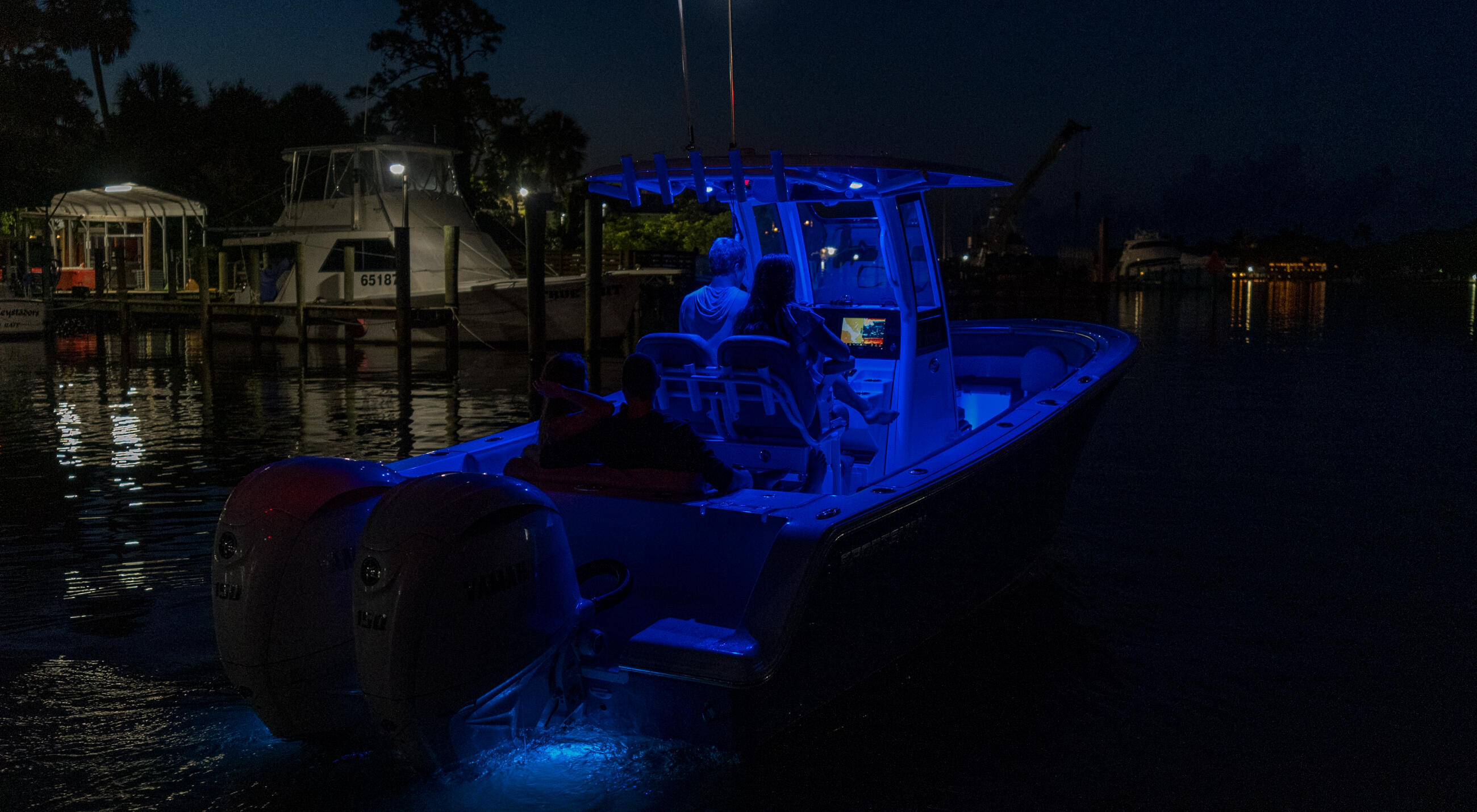 Blue-lit center console fishing boat docked at a marina at night with palm trees and lit dockside structures in background