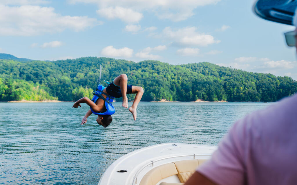 Child doing a backflip off a boat into a forested lake while family watches from the boat deck