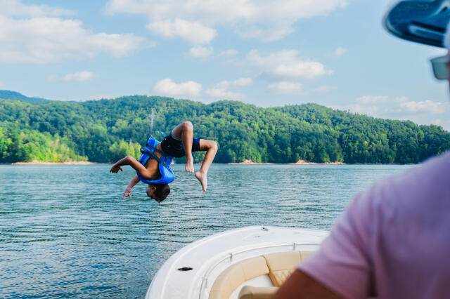Child doing a backflip off a boat into a forested lake while family watches from the boat deck