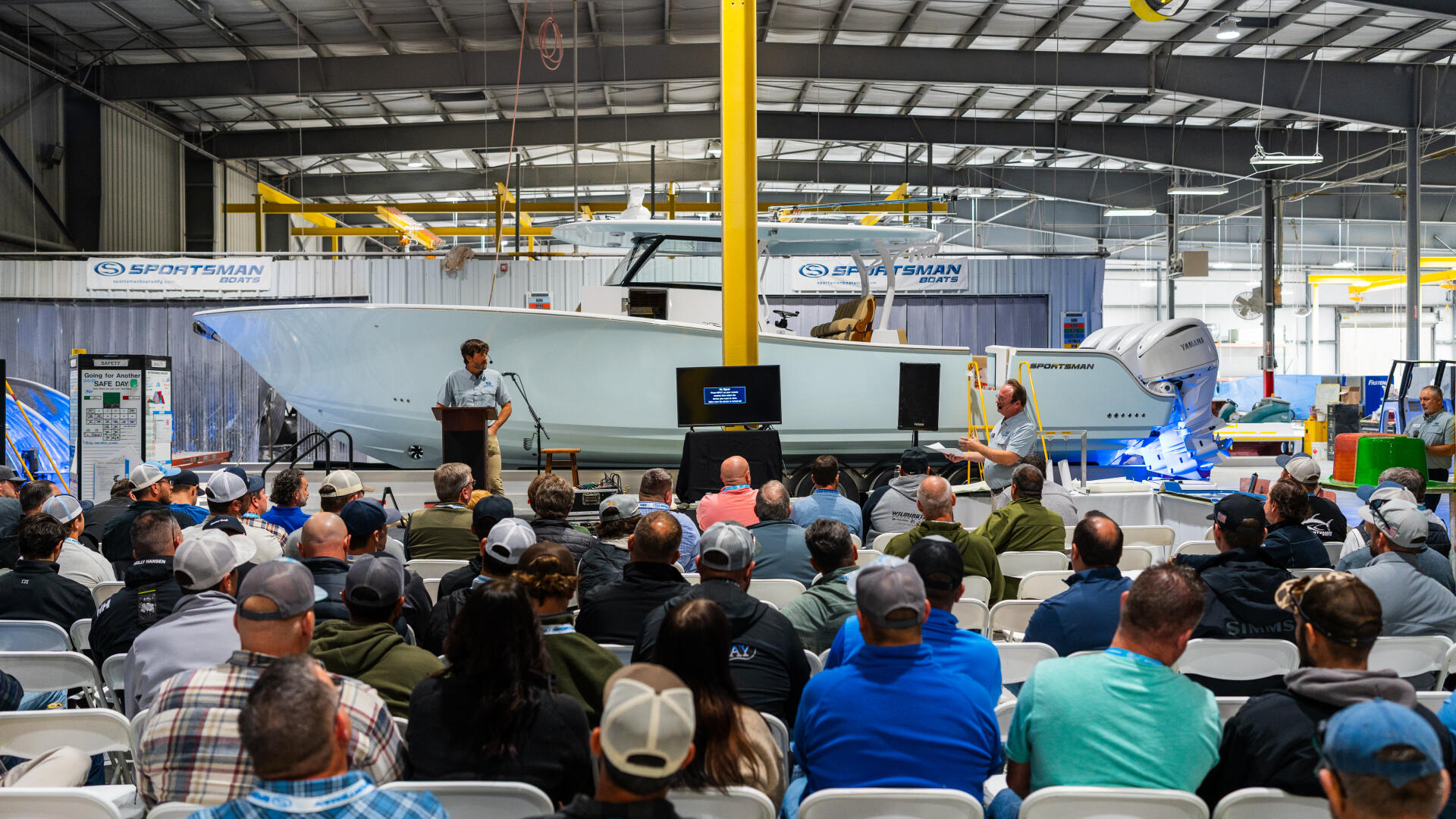 Large crowd seated in manufacturing facility watching presentation next to Sportsman boat on display