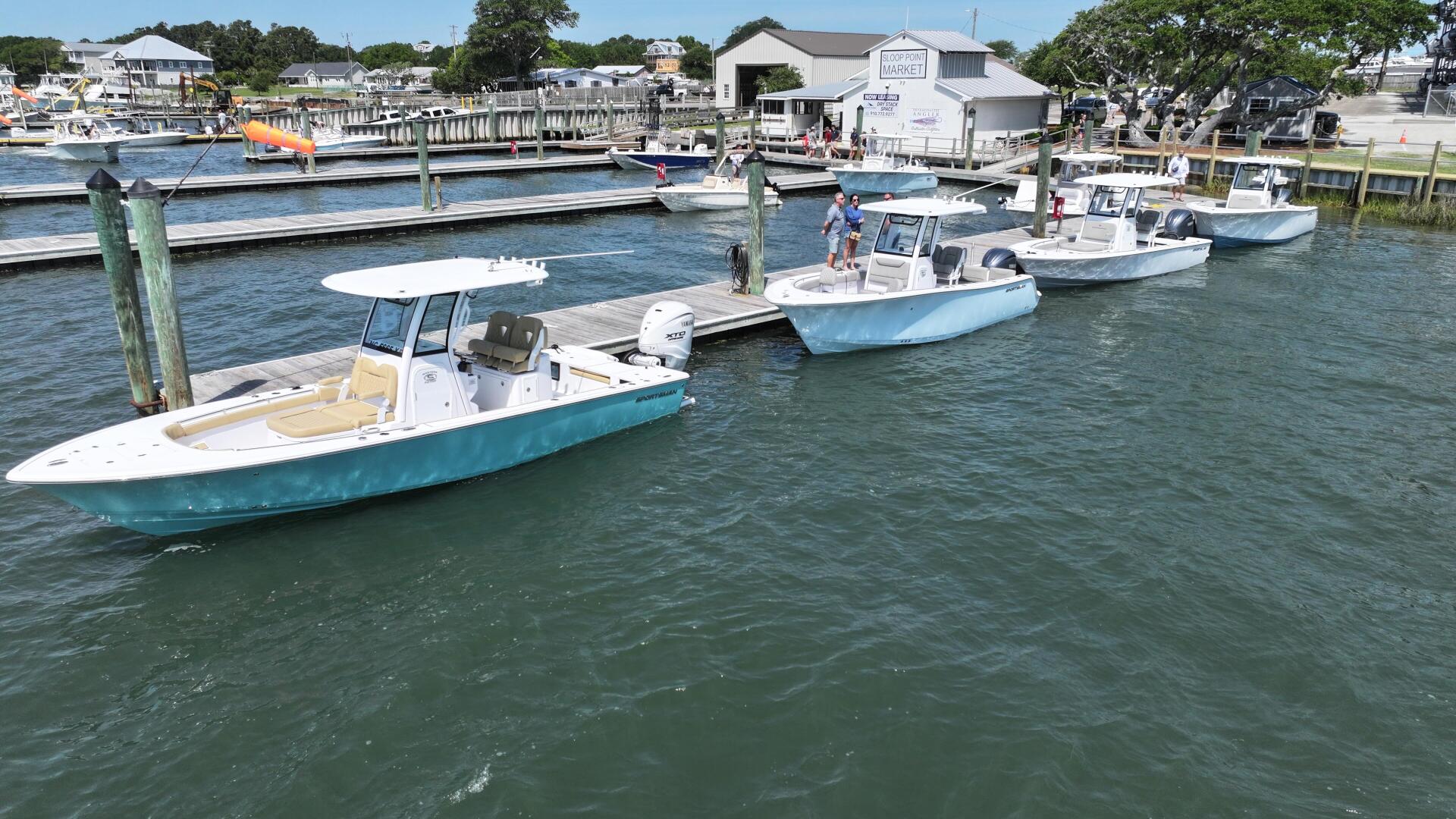 Moored fishing boats with white and turquoise hulls at a busy marina with dockside buildings and green trees