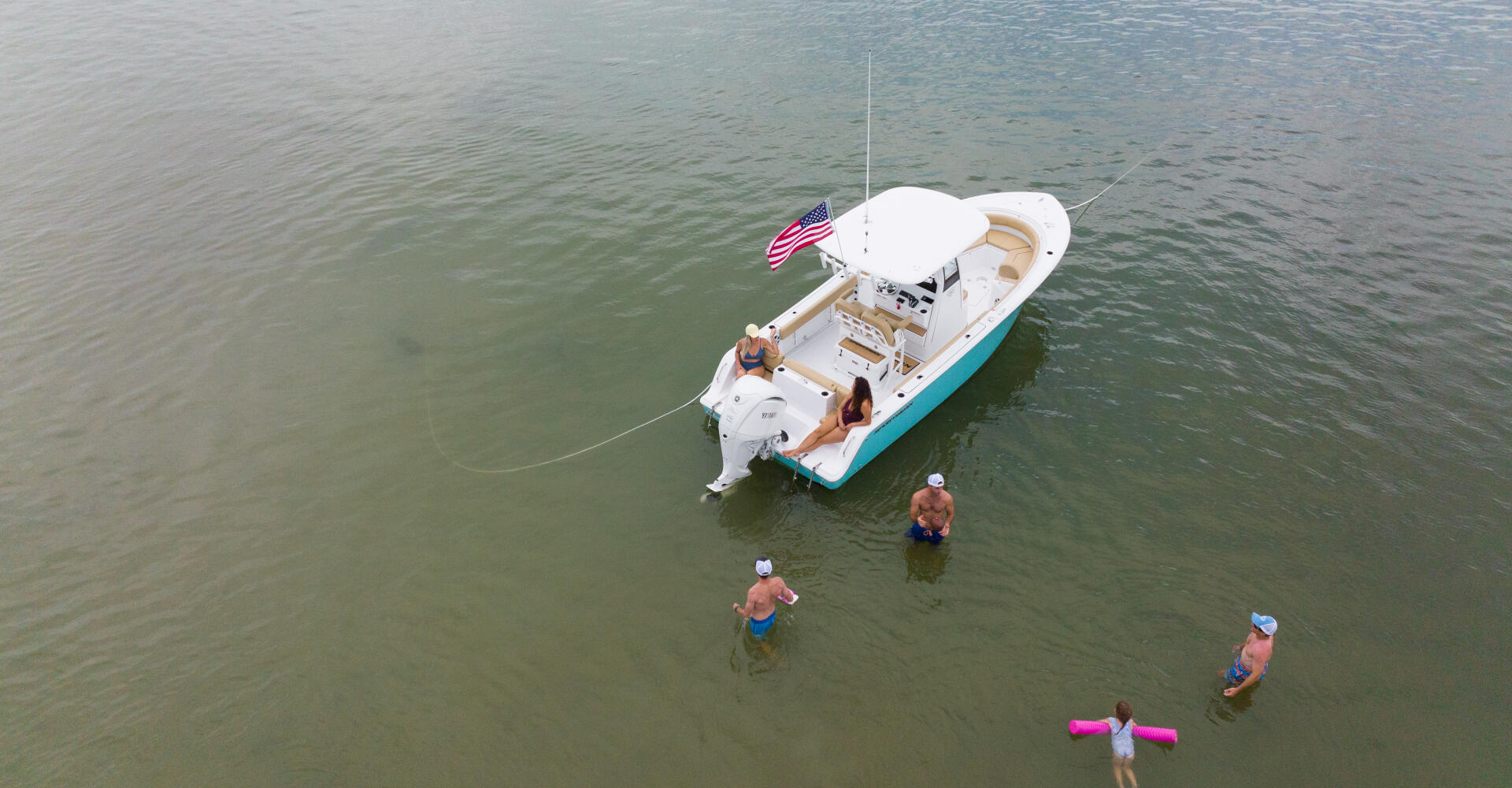 Family swimming and playing in shallow water around a white and turquoise center console boat anchored at a sandbar