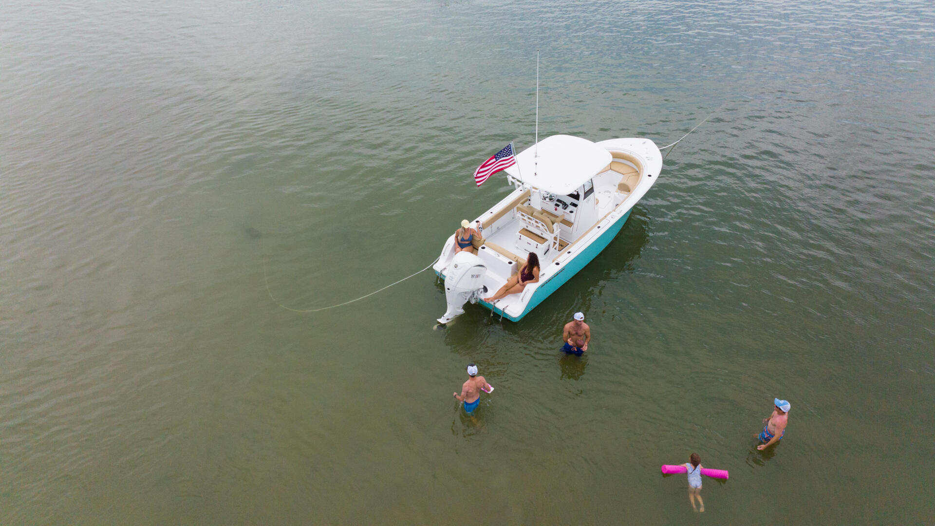 Family swimming and playing in shallow water around a white and turquoise center console boat anchored at a sandbar