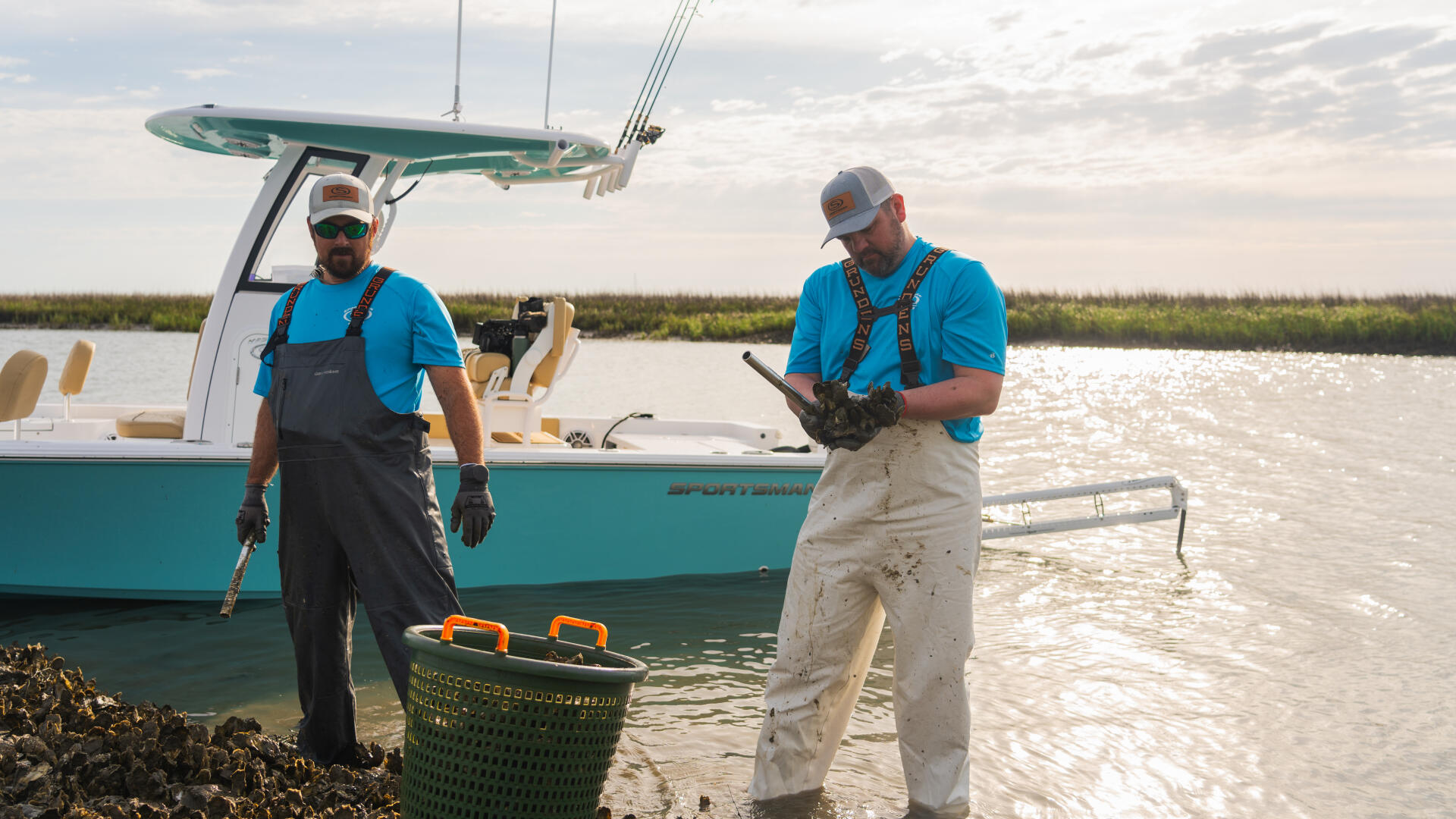 Two men in waders harvest oysters from shallow water near a docked turquoise bay boat