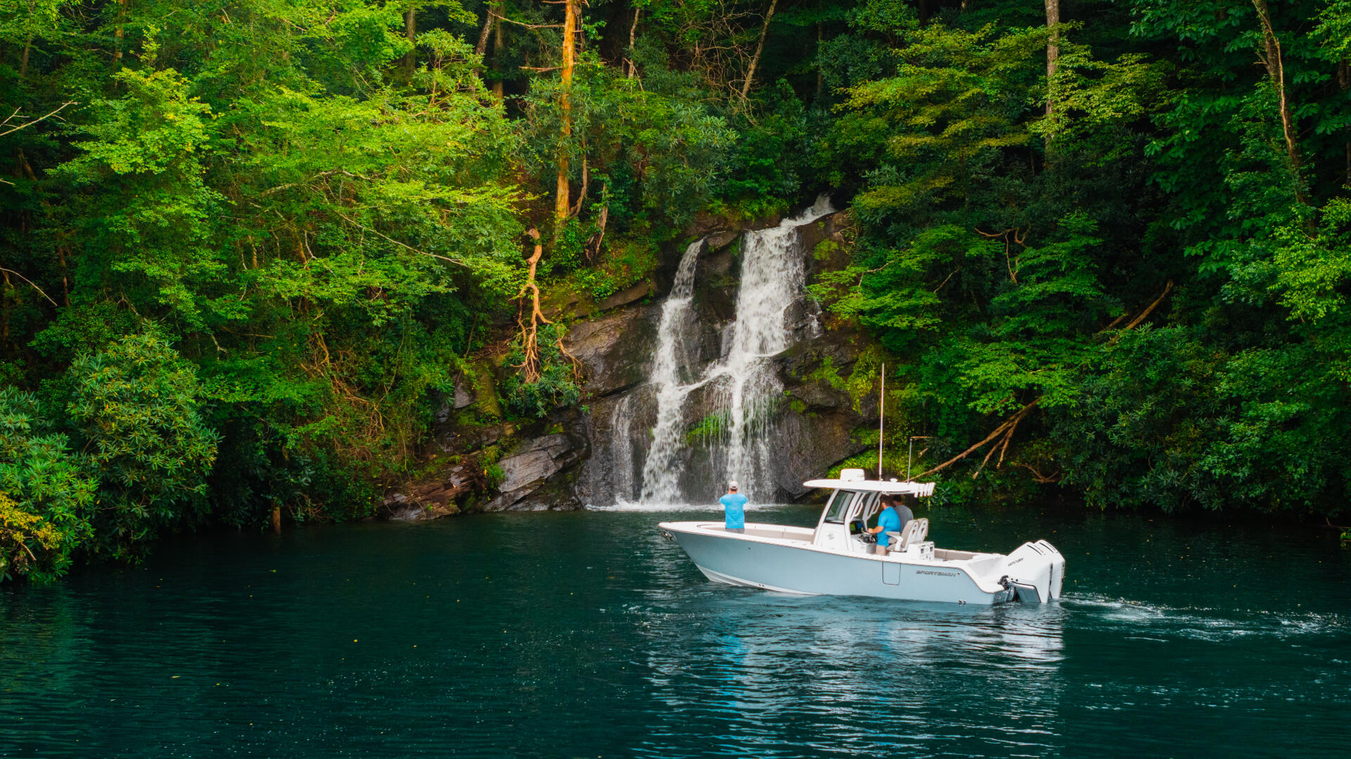 White fishing boat anchored in turquoise water beneath a cascading waterfall surrounded by dense green forest