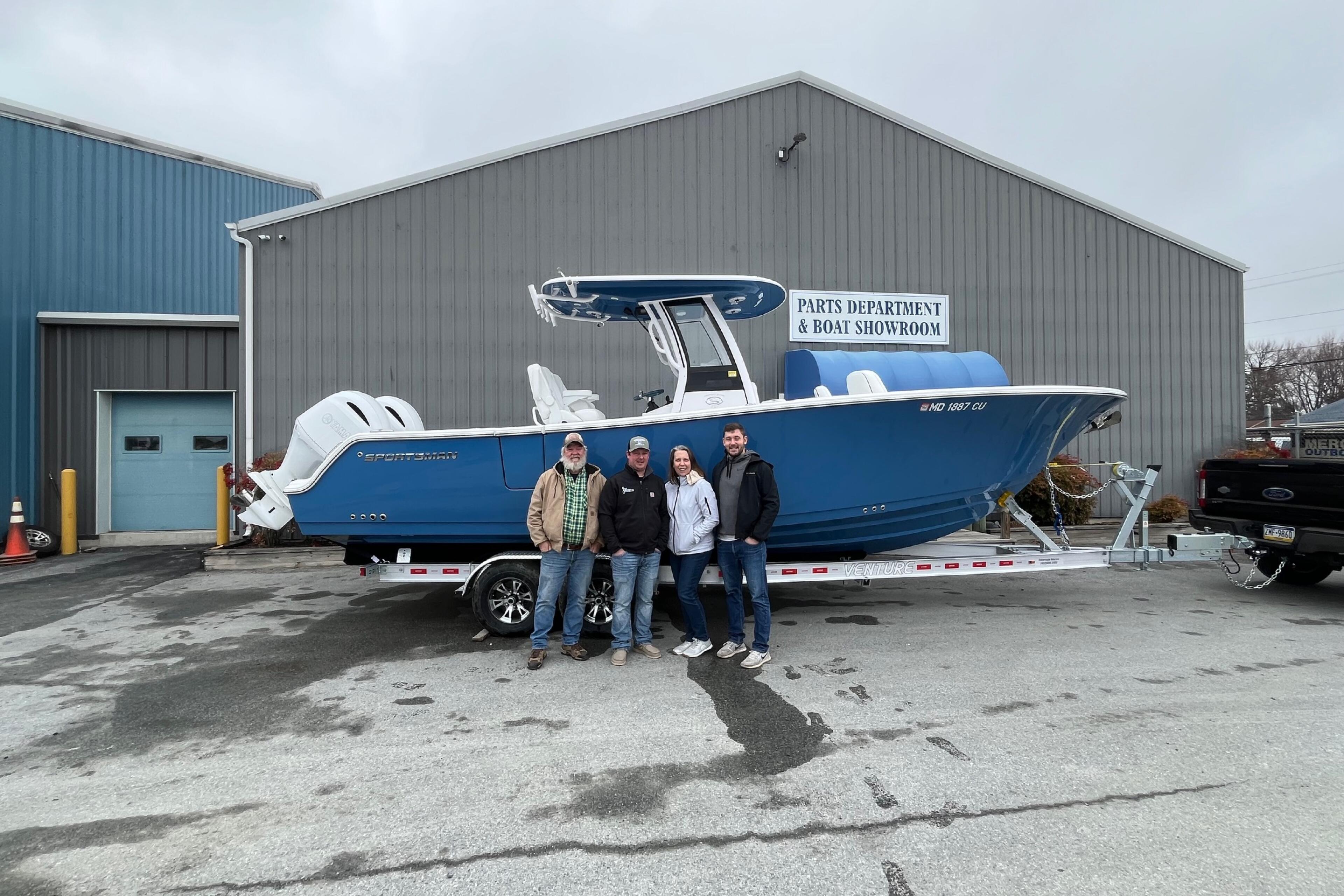 Four people stand in front of a blue sportfishing boat on a trailer at a marine dealership