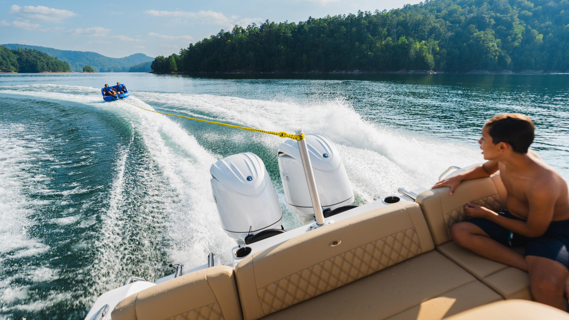 Man relaxing on boat deck while tubing behind a speedboat on scenic mountain lake