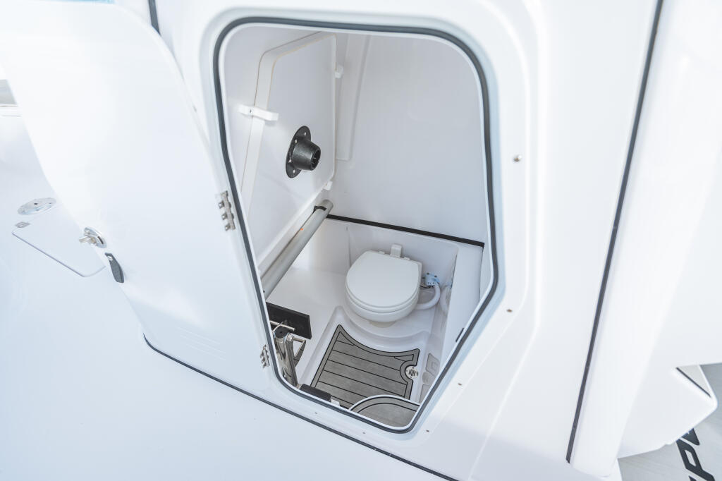 Open cabin door revealing white porcelain toilet and gray floor mat in compact boat head compartment