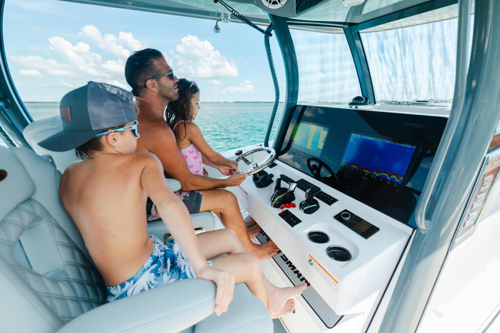Father and children at the helm operating a boat with electronics and controls visible, heading toward turquoise shallow waters