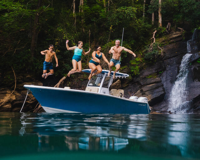 Group of friends jumping off a blue boat into a pristine forest lake with a waterfall in the background