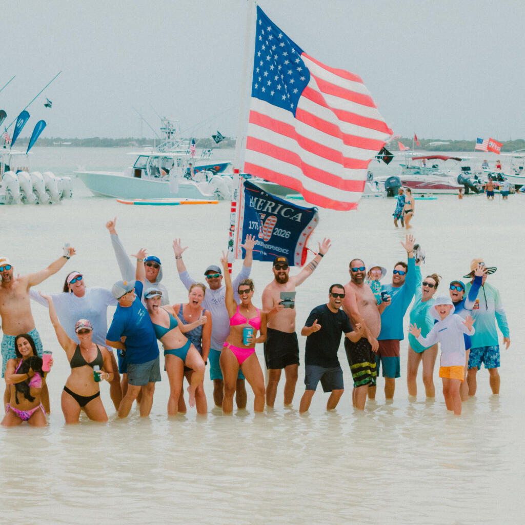 Large group celebrating in shallow beach water with American flag and fishing boats in background