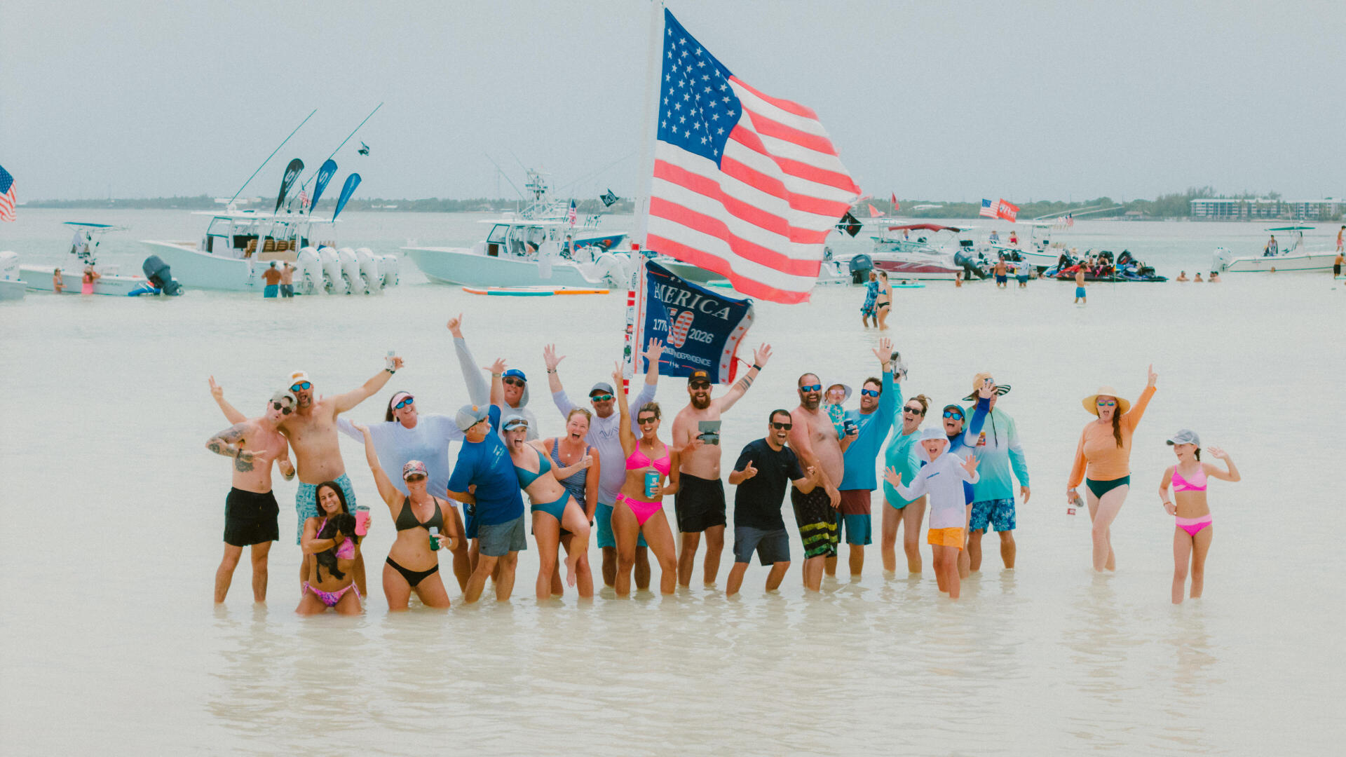 Large group celebrating in shallow beach water with American flag and fishing boats in background