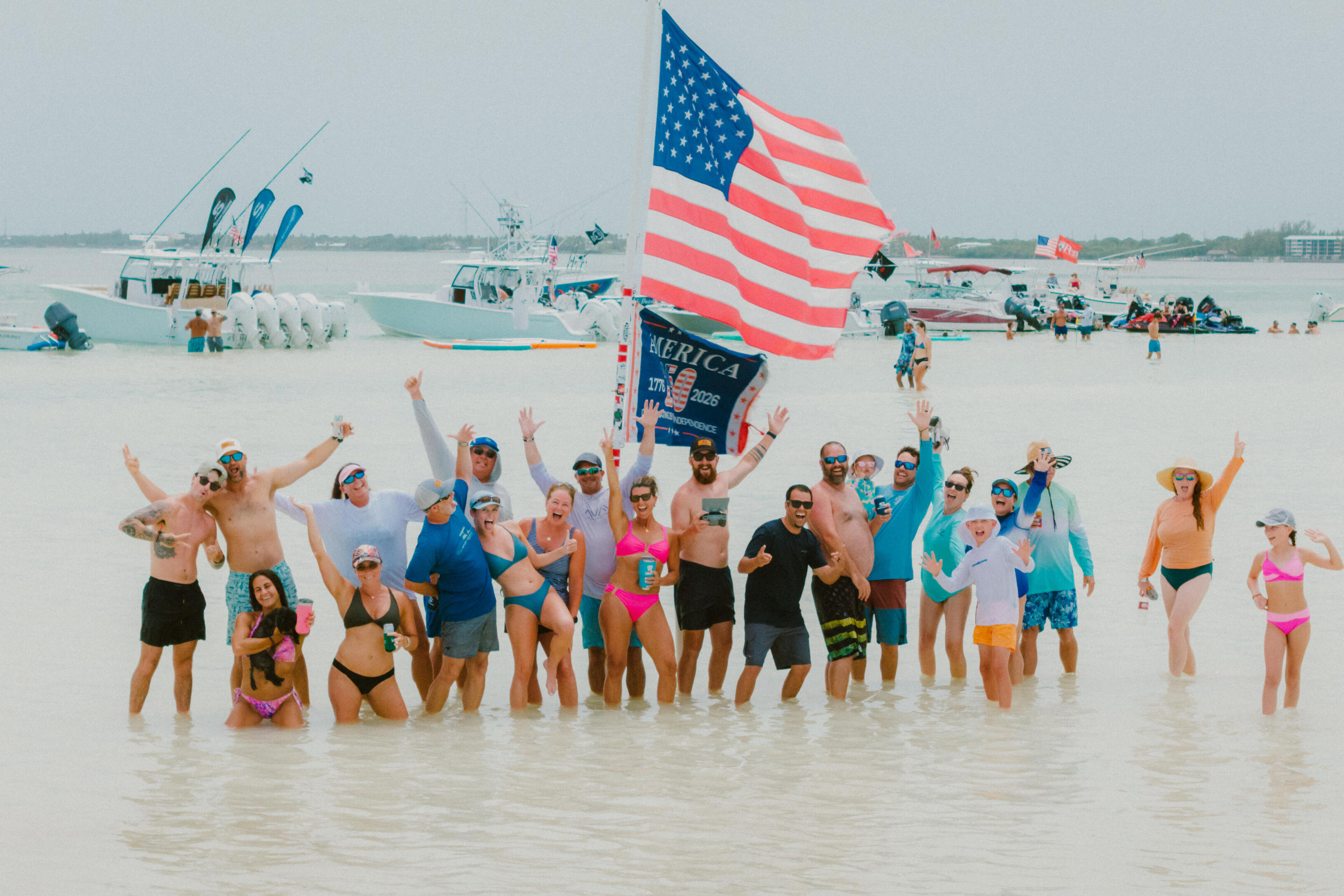 Large group celebrating in shallow beach water with American flag and fishing boats in background