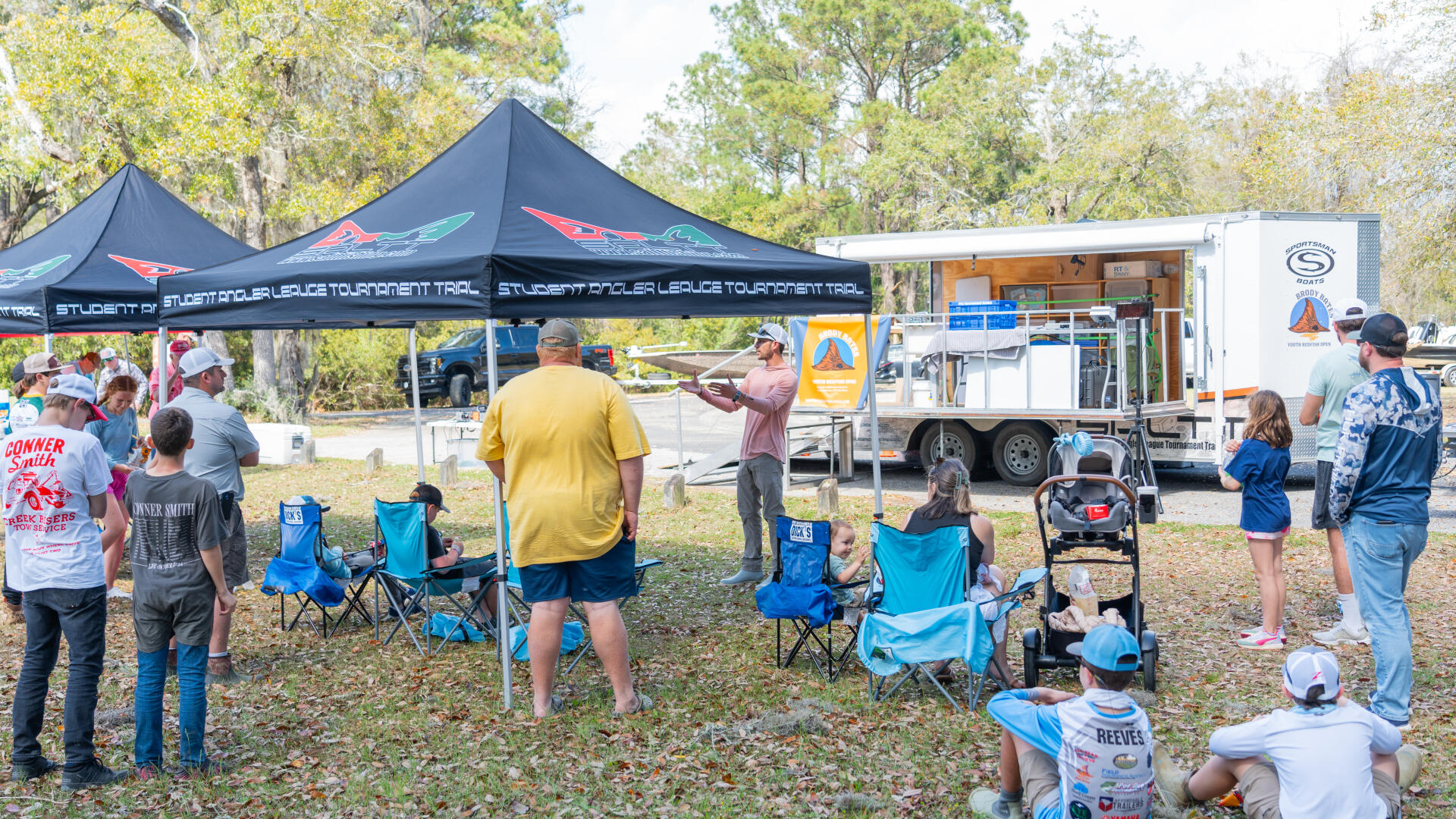 Families and kids gather under a tent at a fishing tournament event with a mobile command center trailer nearby