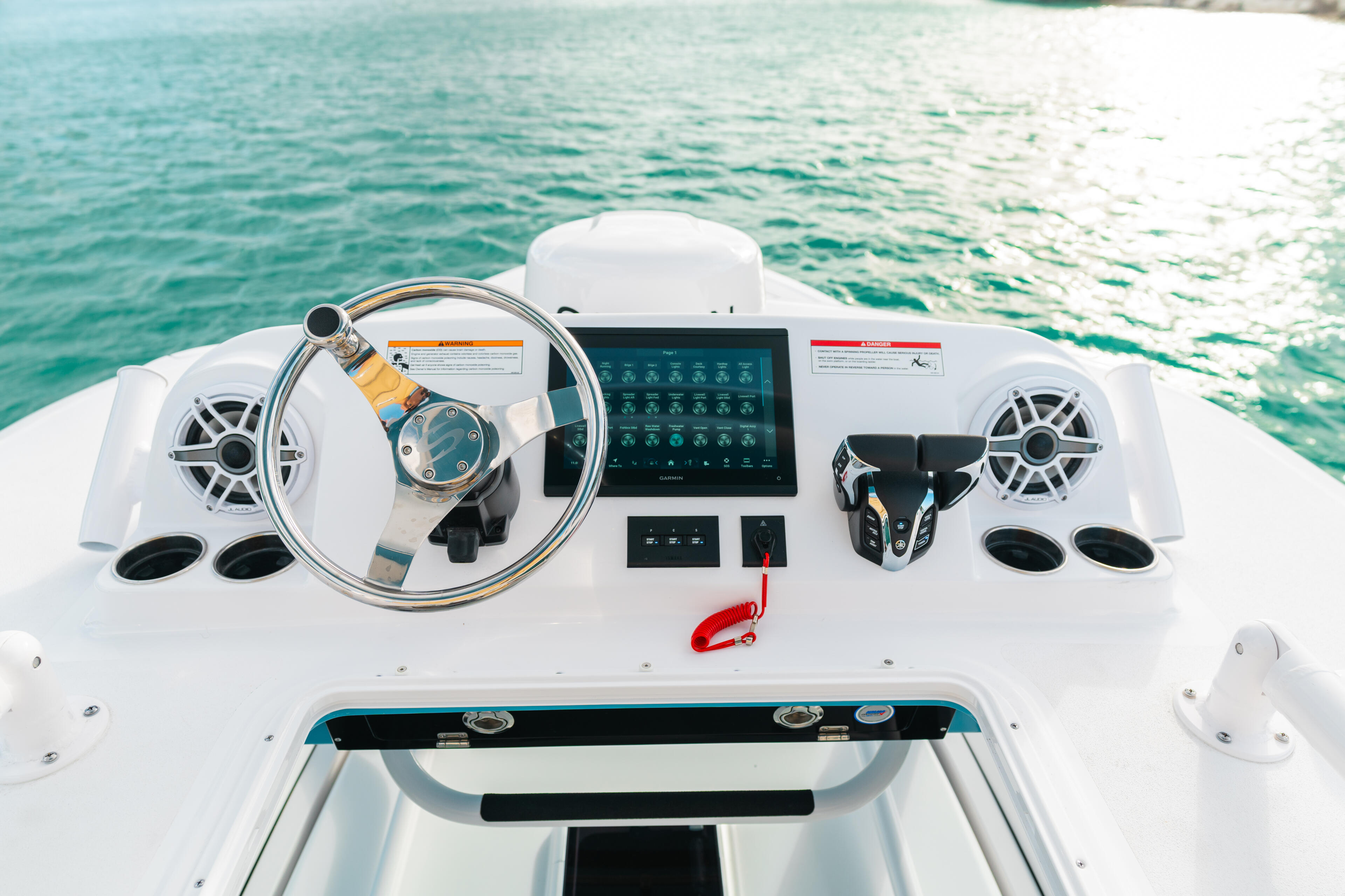 Overhead view of boat helm with steering wheel, touchscreen display, audio speakers, and control panels overlooking turquoise water