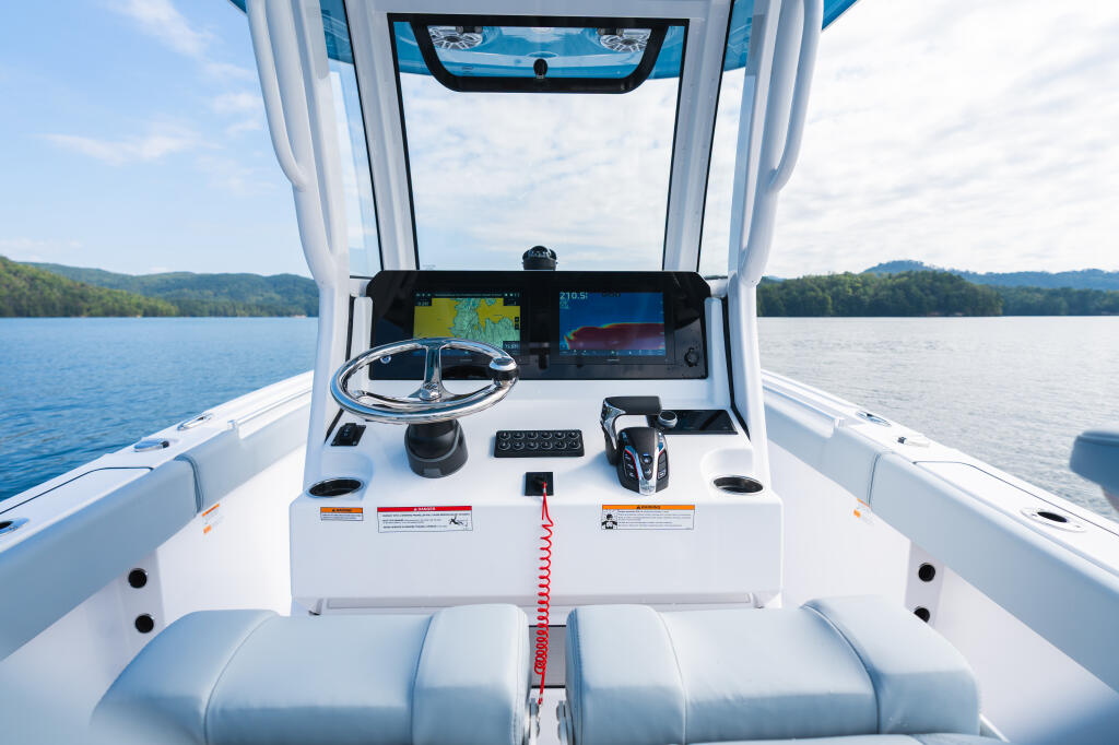 Center console helm with dual touchscreen displays, steering wheel, and control panel overlooking a calm lake with forested shores