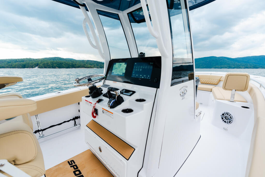 Helm station with dual displays and controls under hardtop, cream seating visible alongside calm lake and forested shoreline