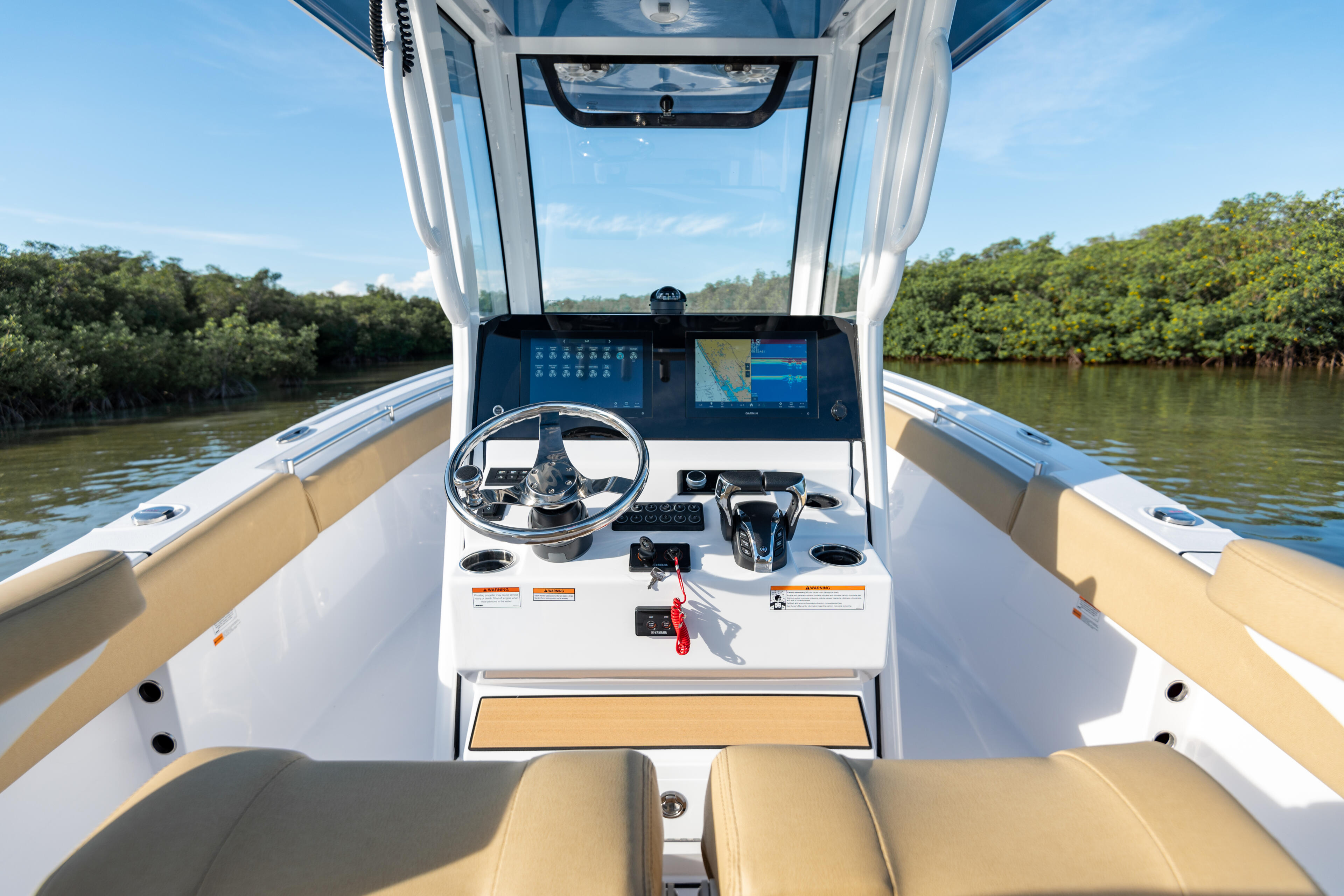 Helm station with dual monitors, steering wheel, and tan seating facing a mangrove shoreline in calm water