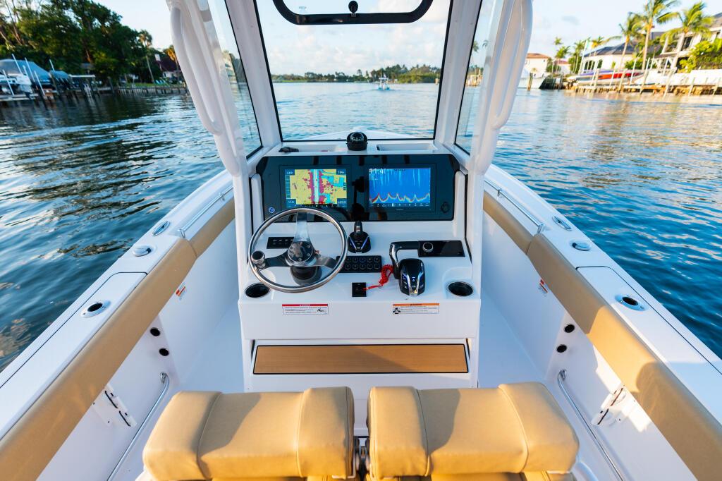Center console helm with dual monitors, steering wheel, and tan cushioned seating overlooking calm waterway