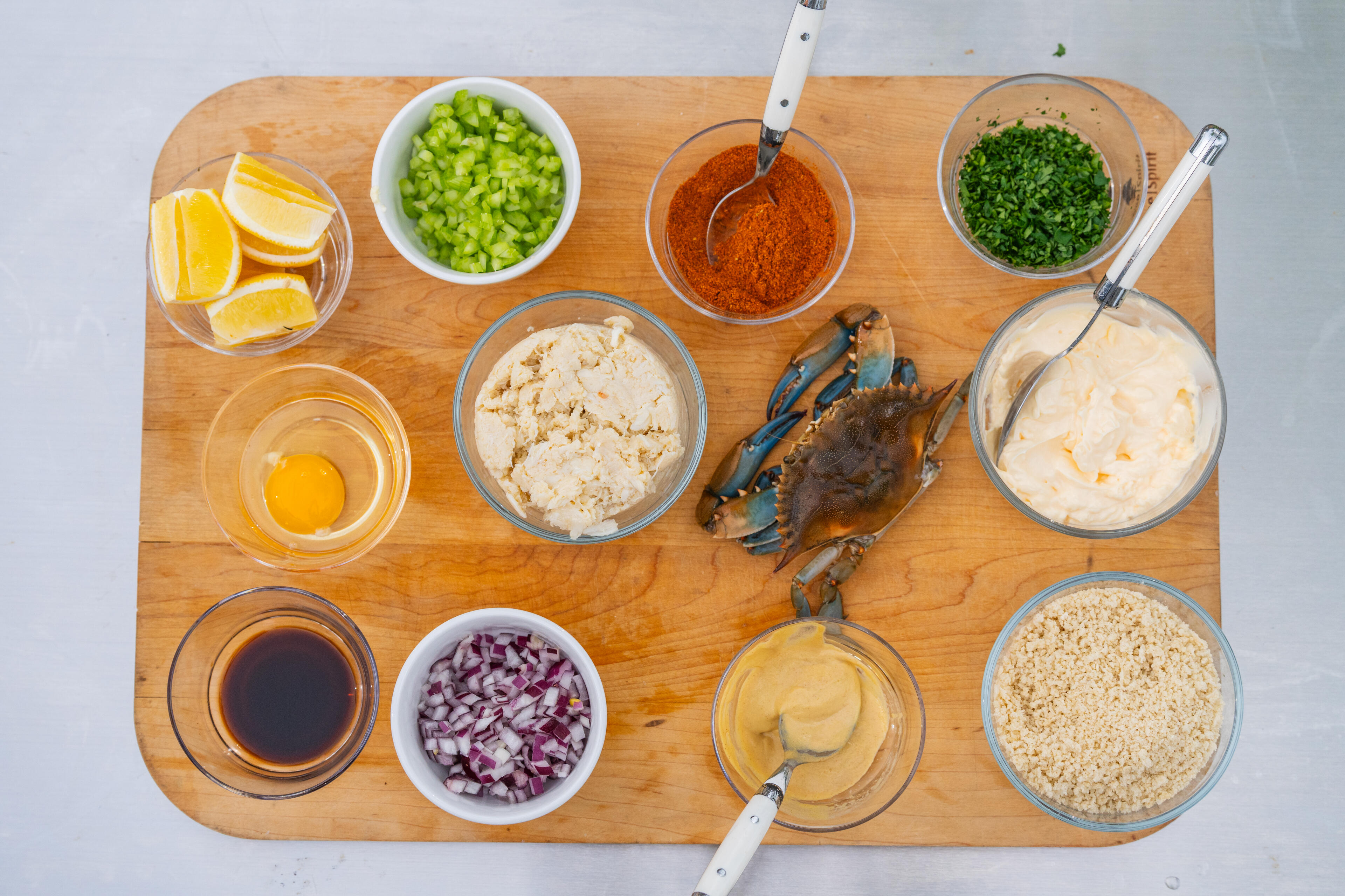 Overhead view of fresh crab cakes and dip ingredients arranged on a wooden board with lemon, celery, spices, and seasonings