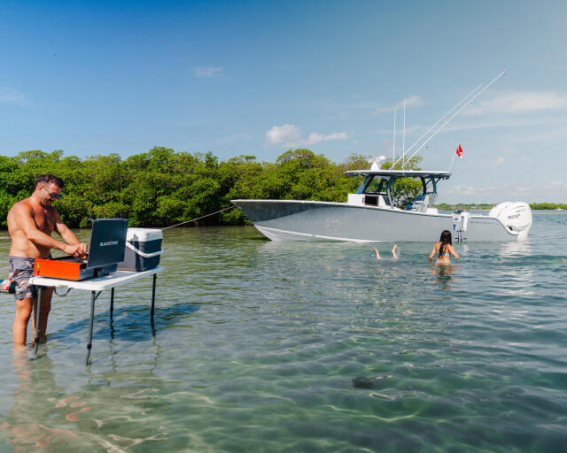 Man at a table in shallow water while families swim near a docked center console boat