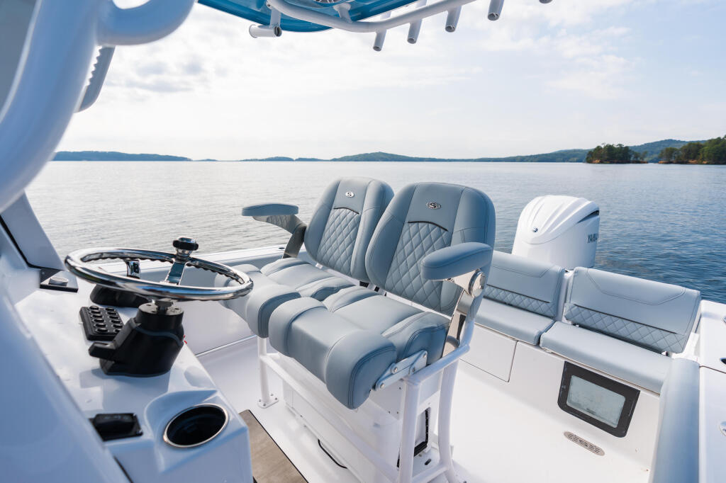 Center console helm with dual blue and white quilted seating, steering wheel, and electronics overlooking calm lake