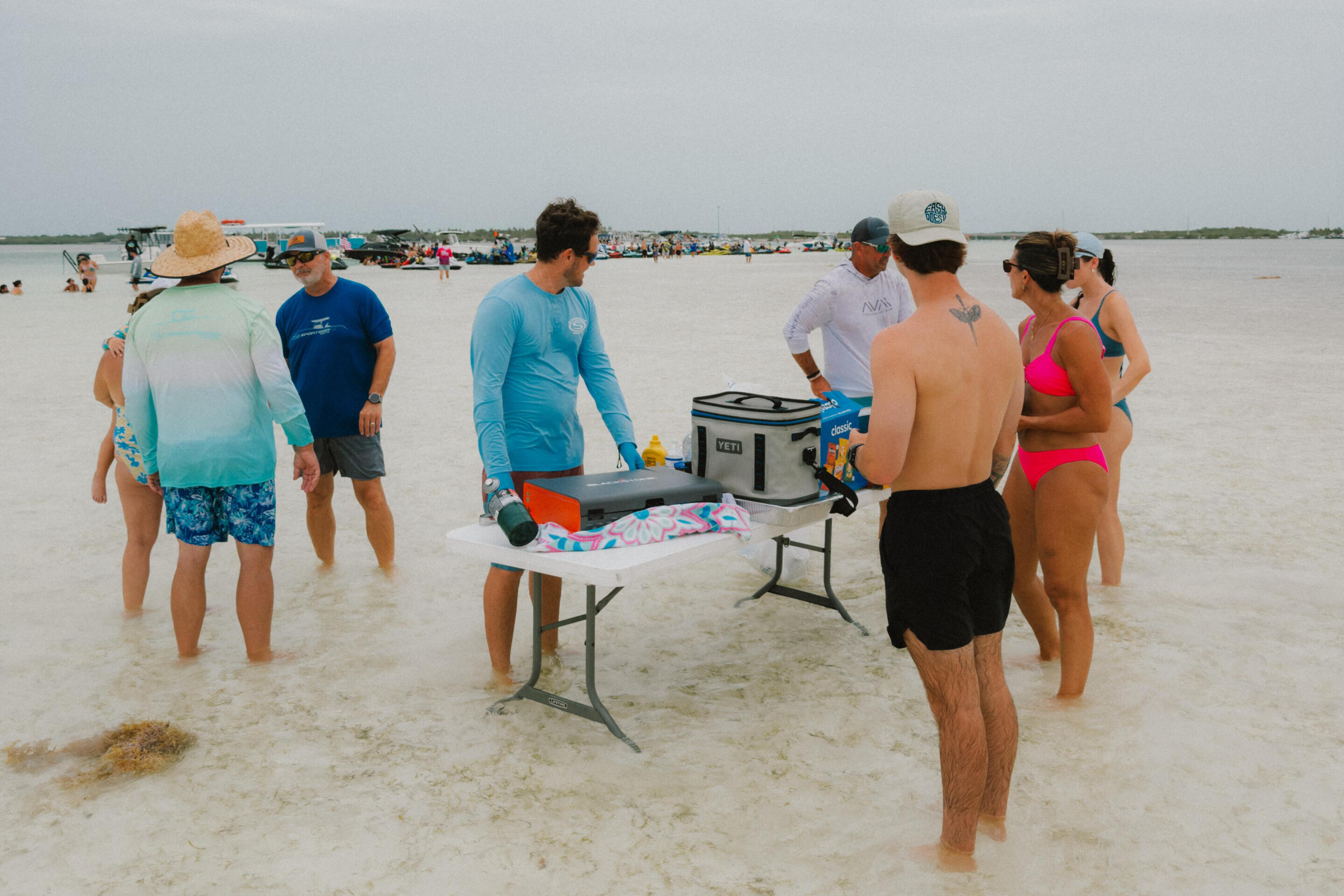 Group of friends gathered around a table on a shallow sandbar beach with coolers and beach gear