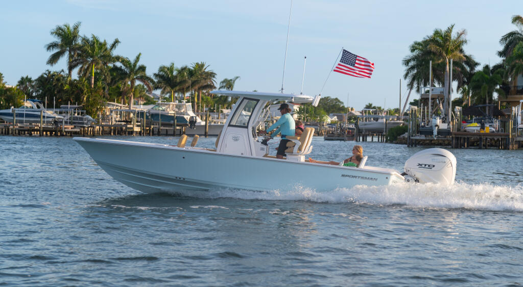 Light blue boat running in shallow water with passengers and American flag, palm trees and marina visible in background