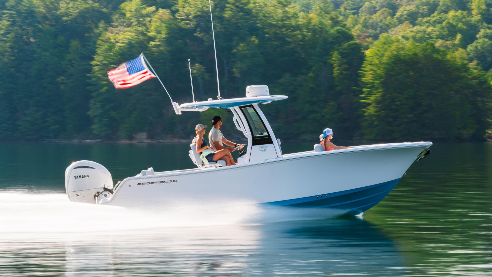Boaters cruising past forested shoreline with American flag flying from hard-top