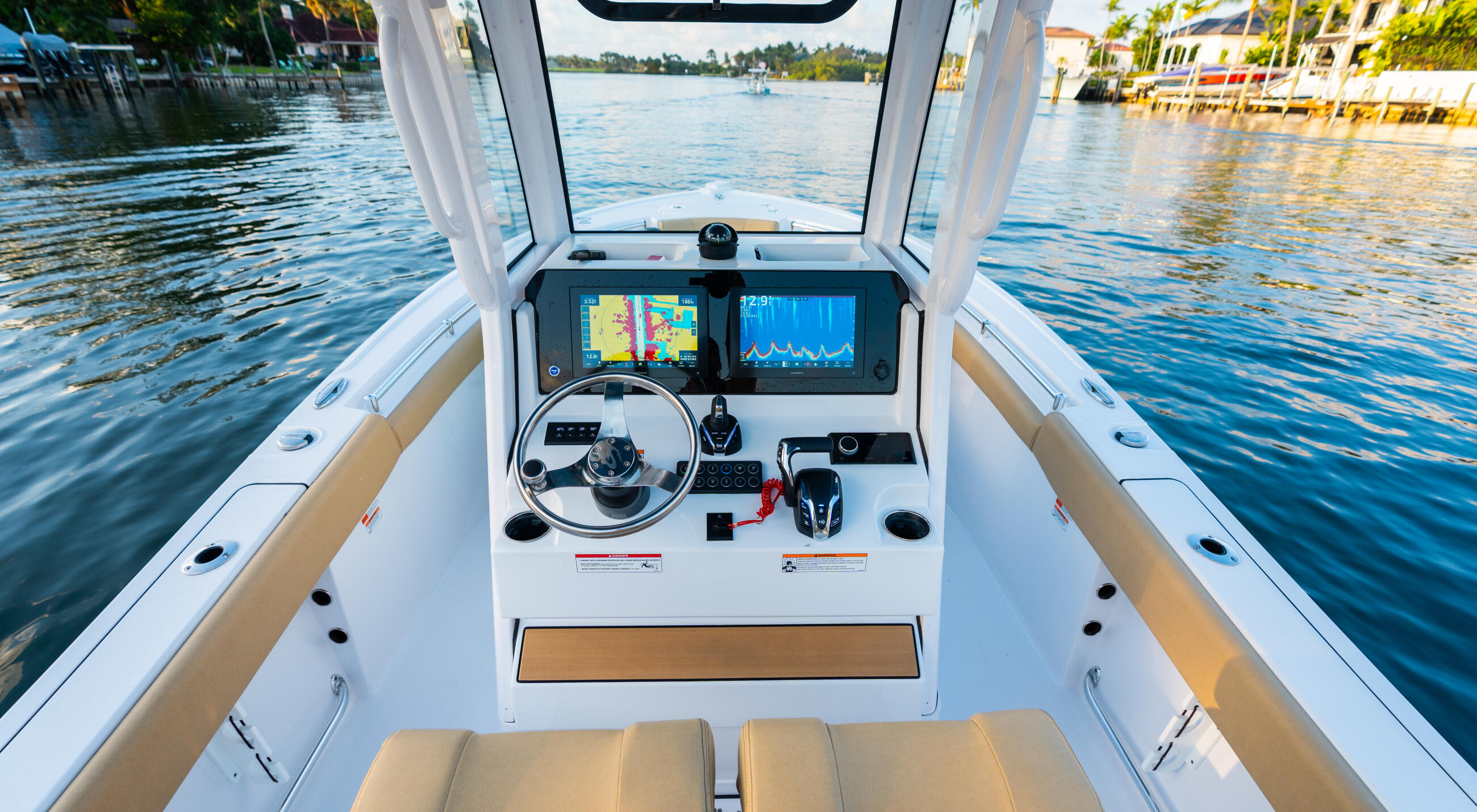 Helm station of center console boat with dual displays, steering wheel, and tan seating facing open water and waterfront homes