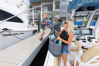 Family of three at a marina boat dock, mother holding a child while father helps lift him onto the boat near the helm station