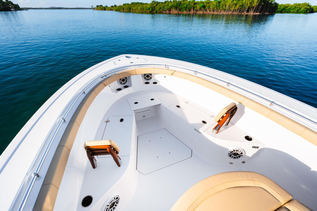 Aerial view of a center console boat's bow with tan and white seating, storage compartments, and mangrove-lined shoreline beyond