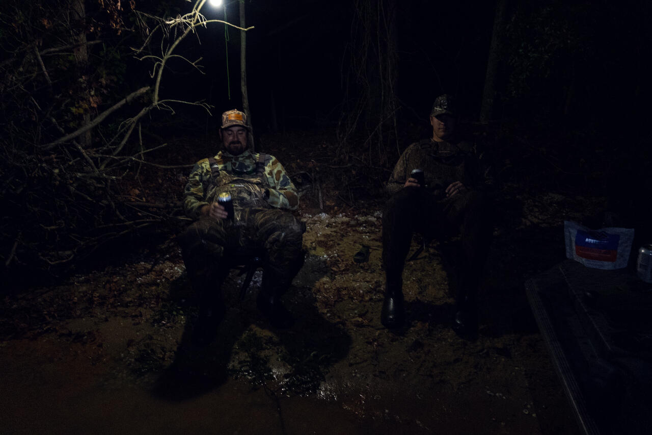 Two anglers in camouflage gear stand at a nighttime campsite with headlamps and a rope strung overhead