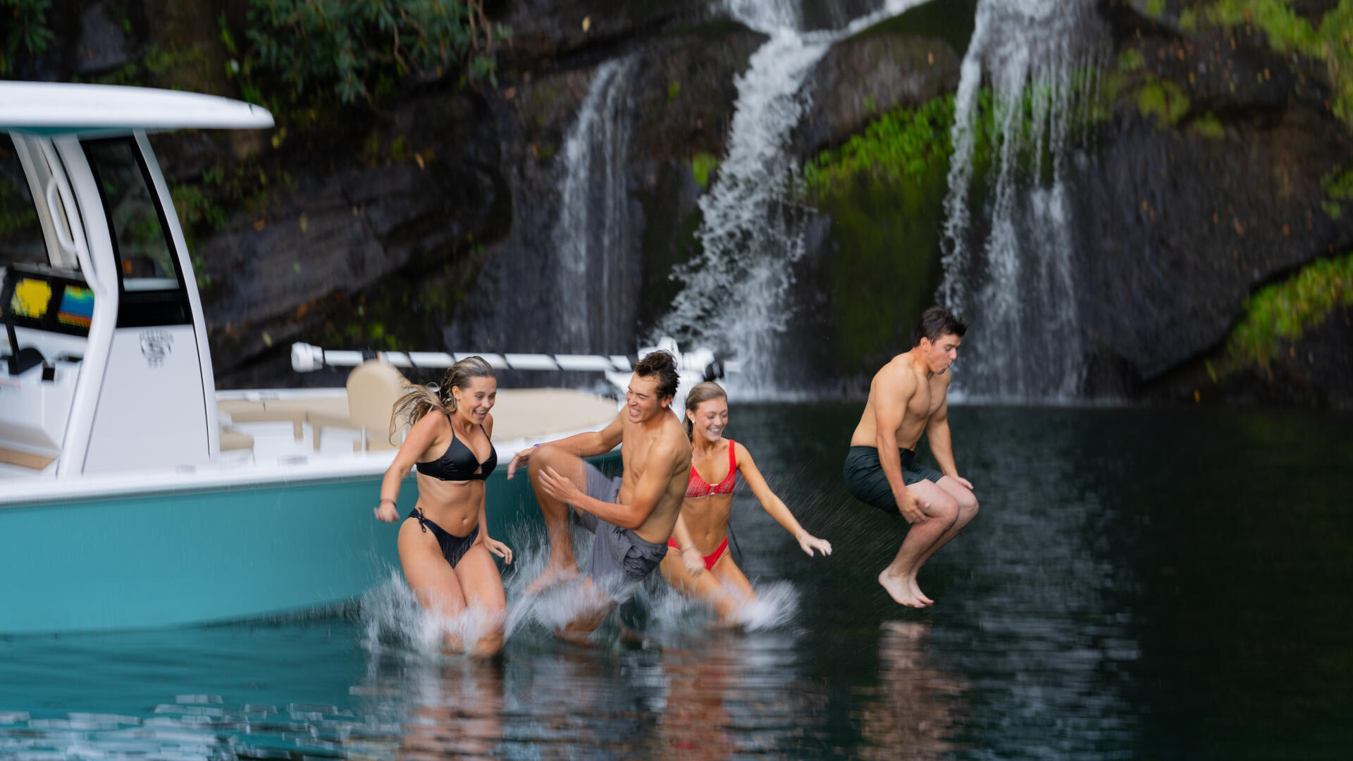 Group of friends swimming and playing in clear water near a boat with a waterfall in the background