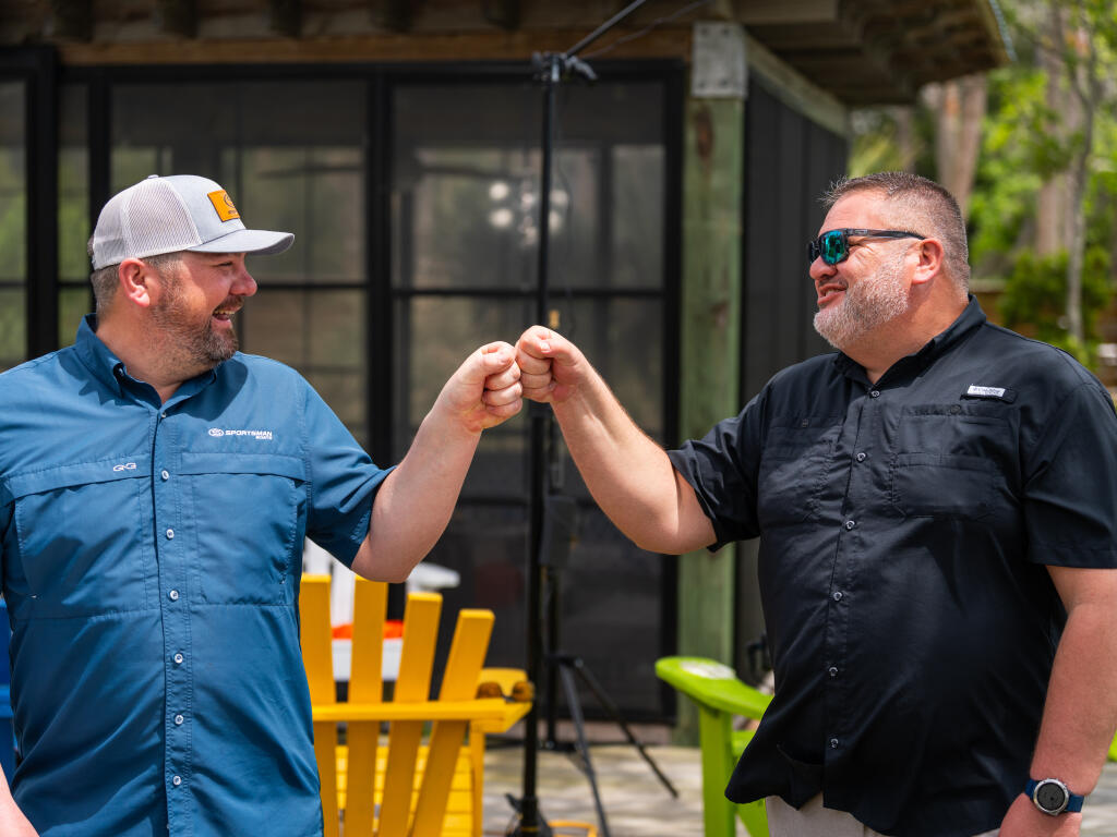 Two men greet each other with a fist bump at a marina, wearing branded work shirts