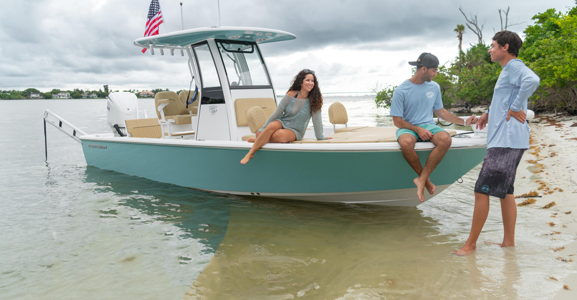 Three people enjoying a turquoise and white center console boat anchored in shallow sandbar waters with a beach shoreline in background