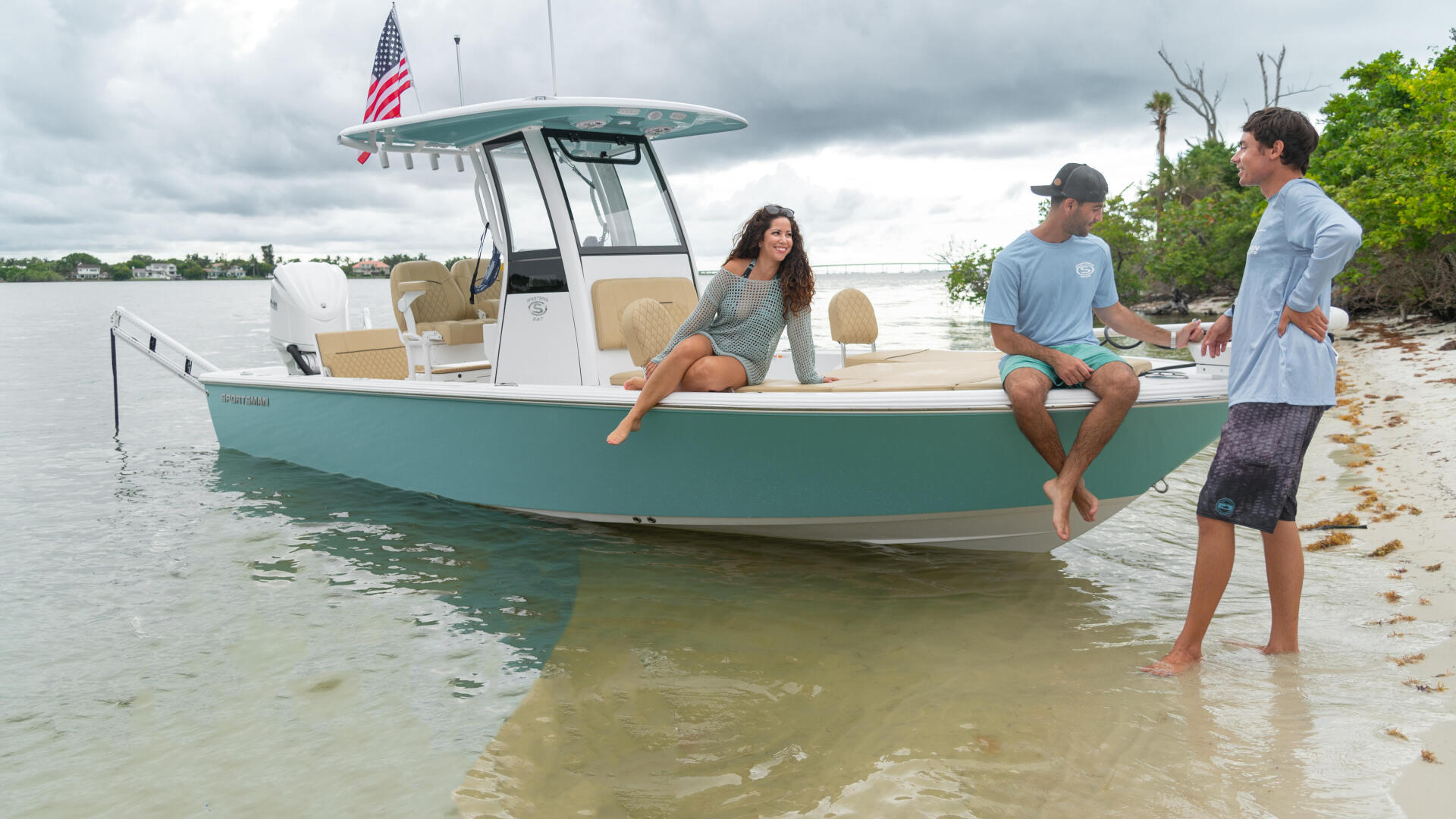 Three people enjoying a turquoise and white center console boat anchored in shallow sandbar waters with a beach shoreline in background