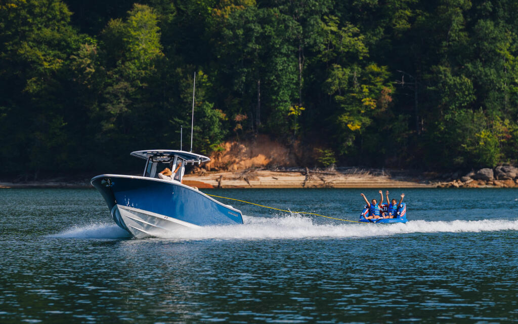 Blue and white boat towing a group of people on a tube across a lake surrounded by forested shoreline