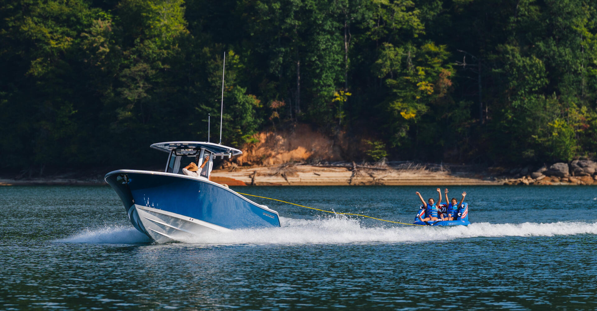 Blue and white boat towing a group of people on a tube across a lake surrounded by forested shoreline