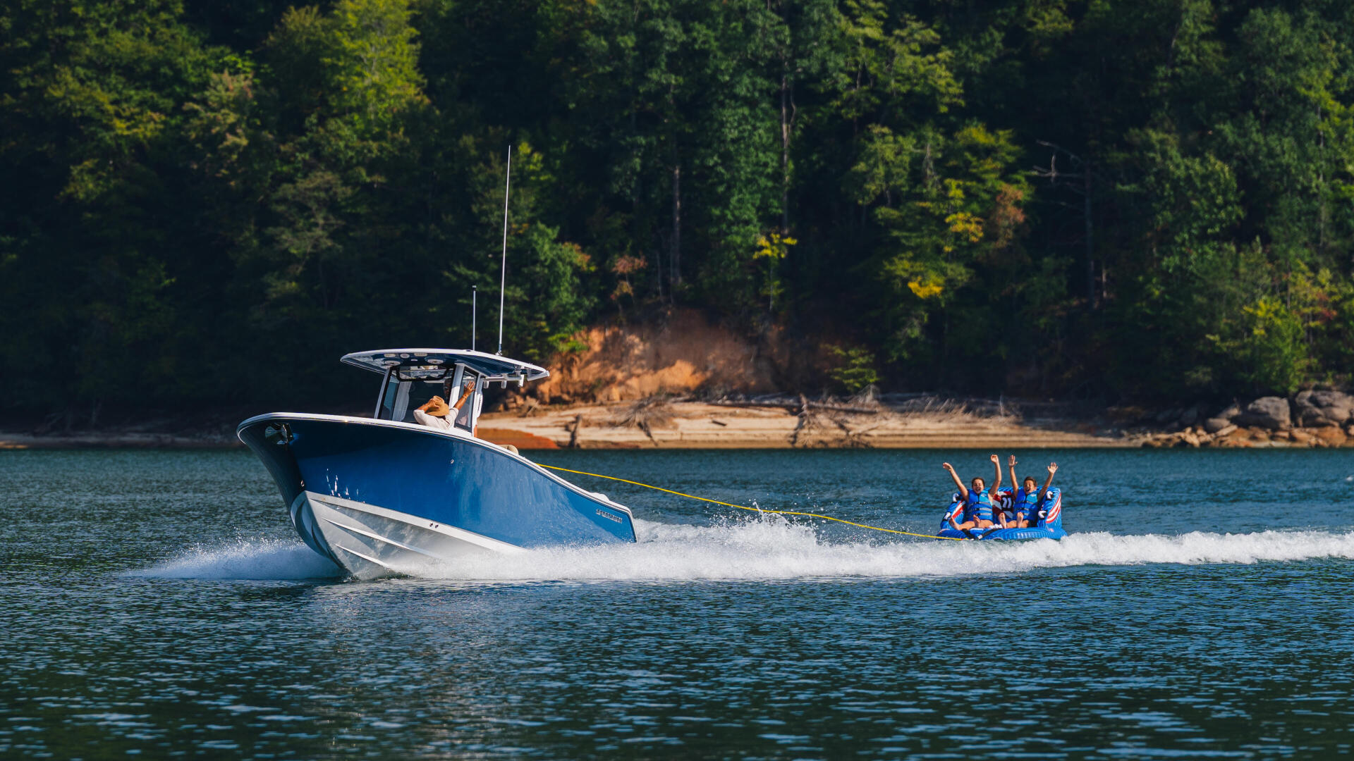 Blue and white boat towing a group of people on a tube across a lake surrounded by forested shoreline