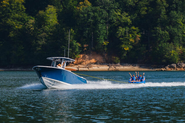Blue and white boat towing a group of people on a tube across a lake surrounded by forested shoreline