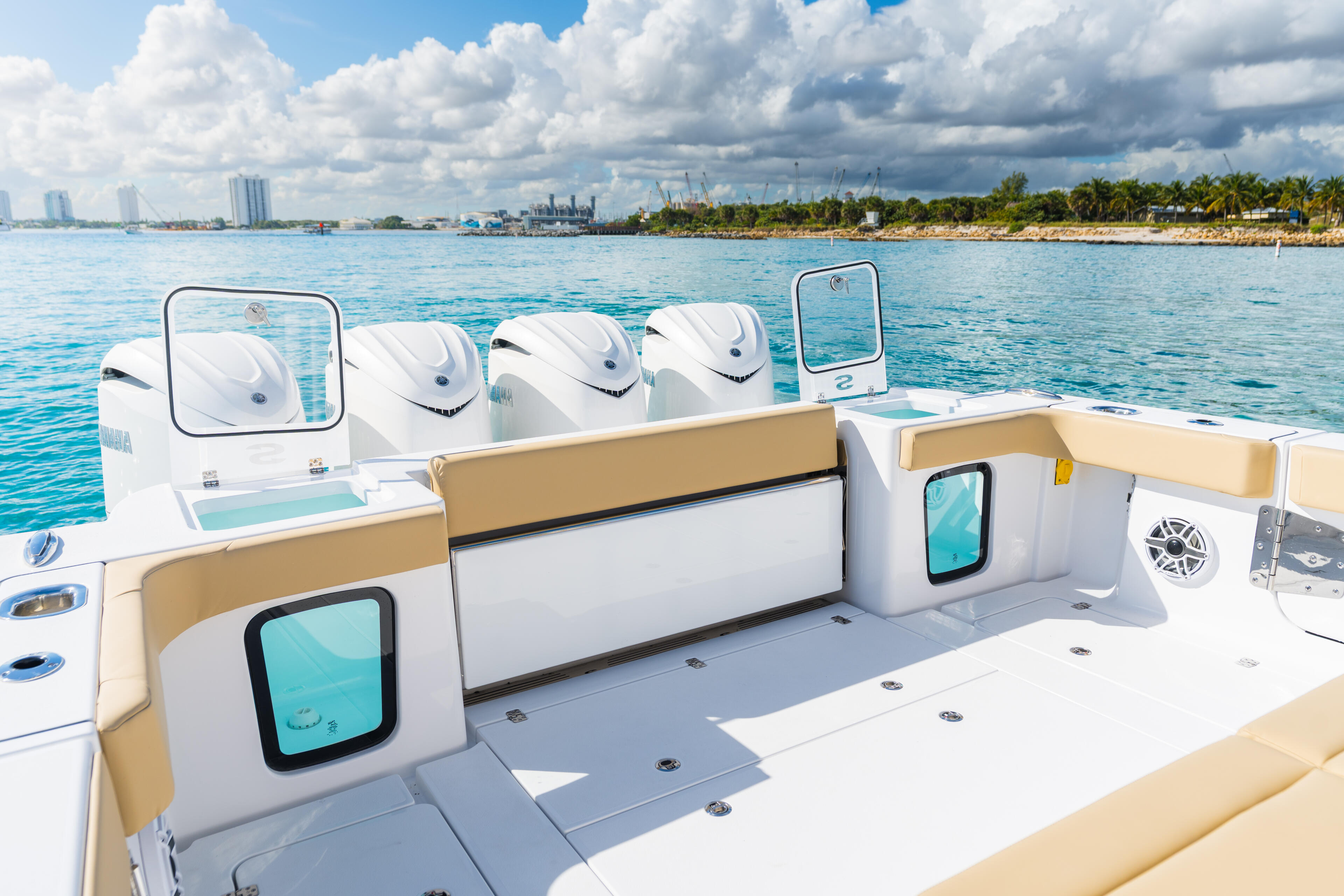 Open deck with tan upholstered seating, white console, and four outboard engines overlooking turquoise water and coastal city skyline