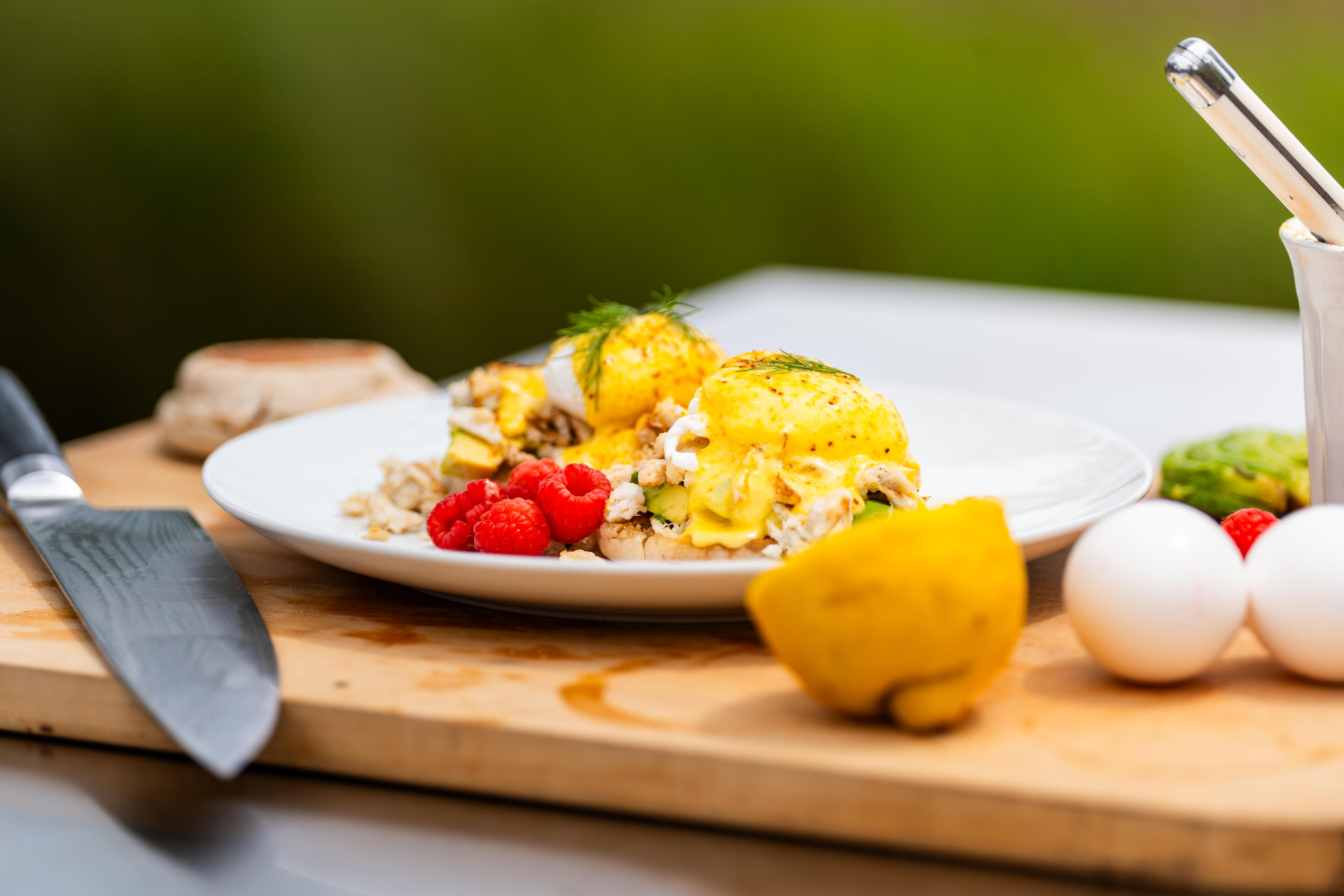 Plated eggs Benedict with hollandaise sauce, fresh raspberries, and garnish on a wooden prep surface with citrus and eggs nearby