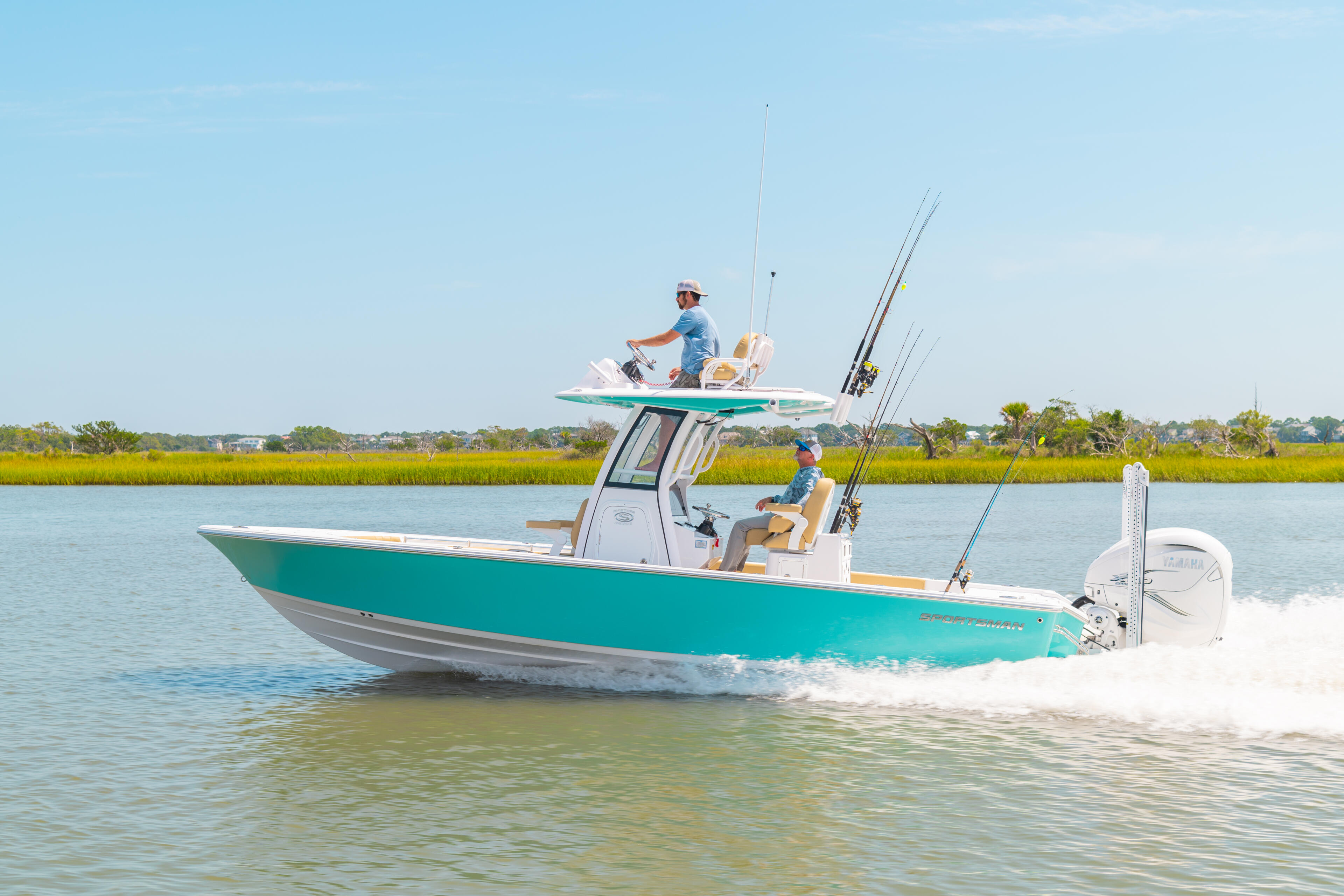 Turquoise and white fishing boat with two anglers and multiple fishing rods running through shallow coastal water near a grassy shoreline