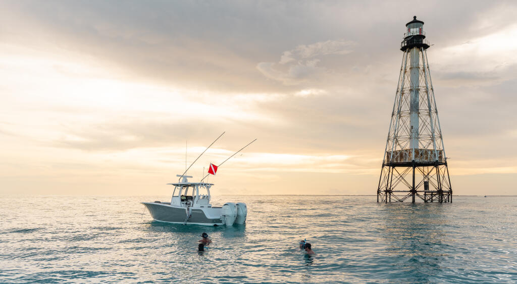 Center console boat anchored offshore near a lighthouse with swimmers in calm water at sunset