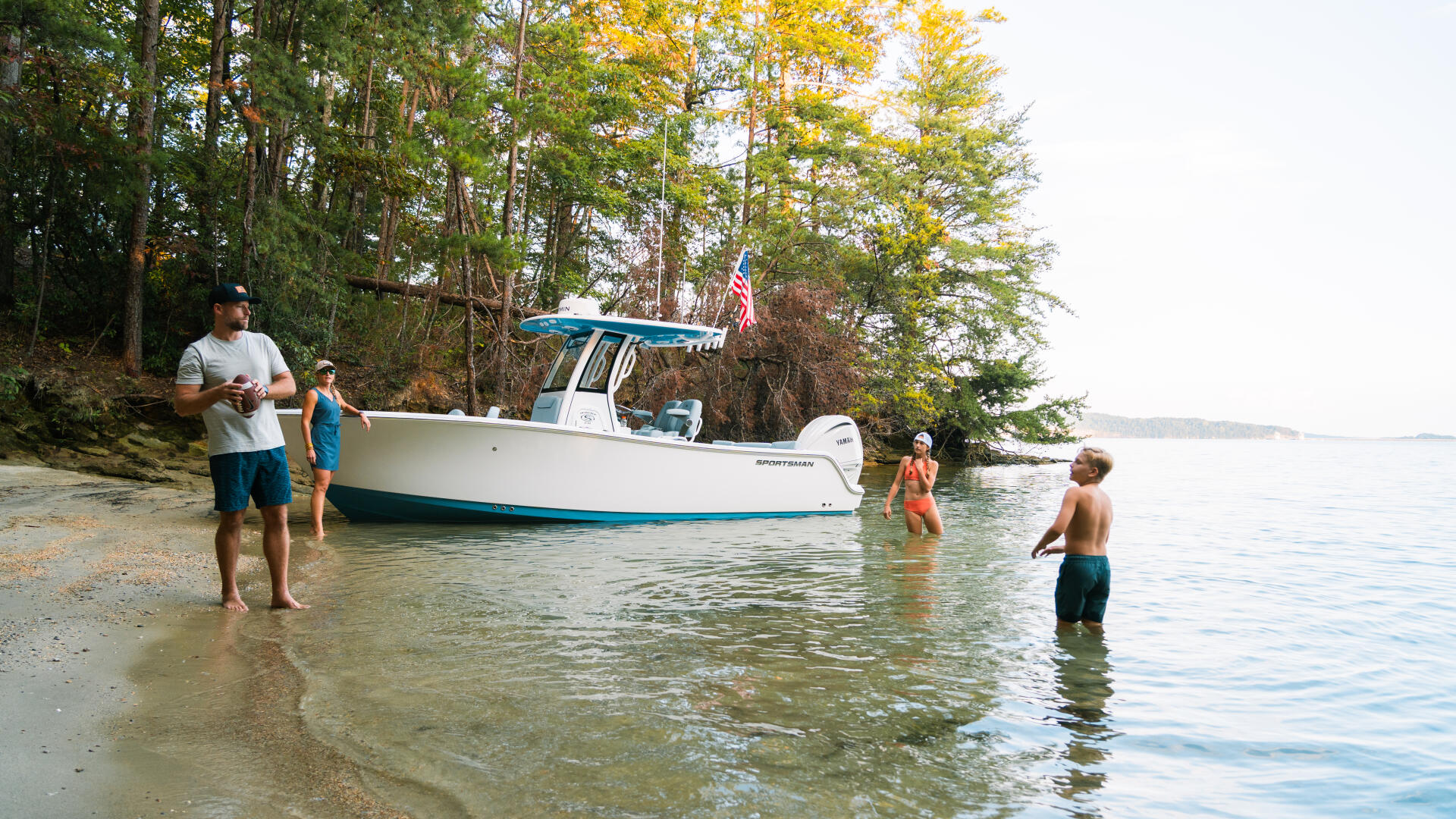 Family swimming and wading in shallow water next to a docked center console boat at a sandy beach cove surrounded by trees