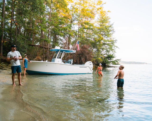 Family swimming and wading in shallow water next to a docked center console boat at a sandy beach cove surrounded by trees