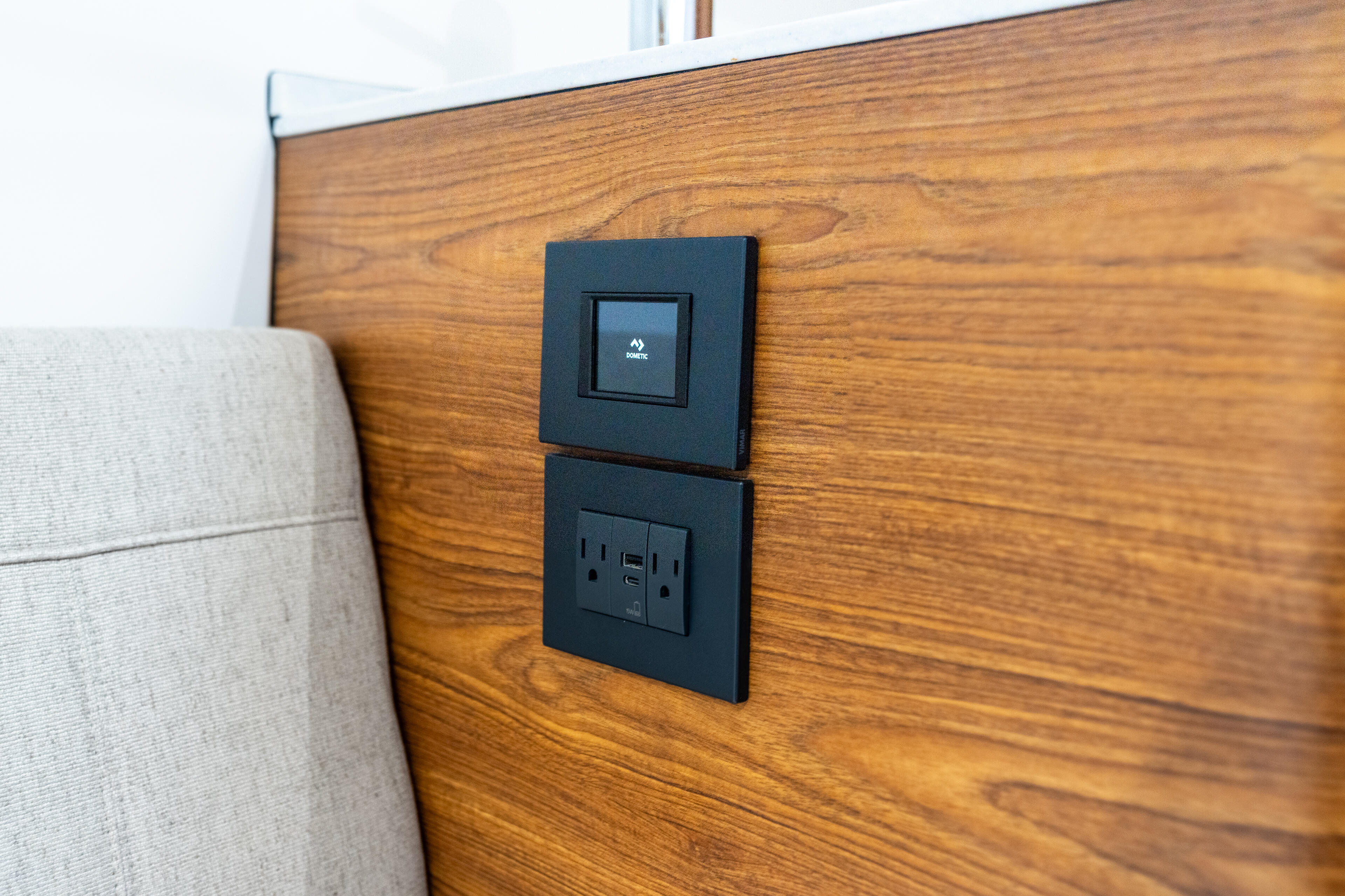 Black smart control panel and dual 110V outlets mounted on wood-grain cabin wall beside upholstered seating