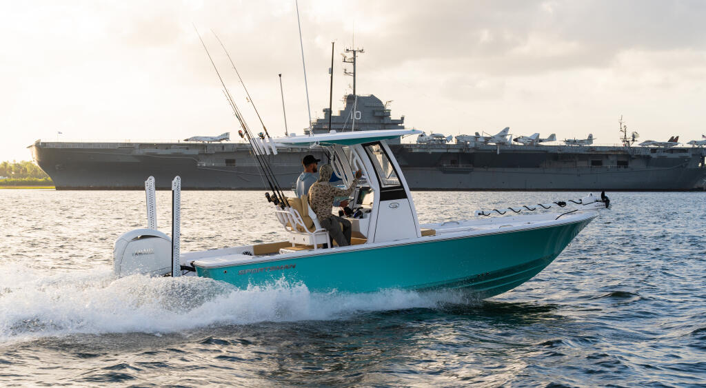 Turquoise and white center console fishing boat running through shallow water with anglers aboard, large navy ship visible in background