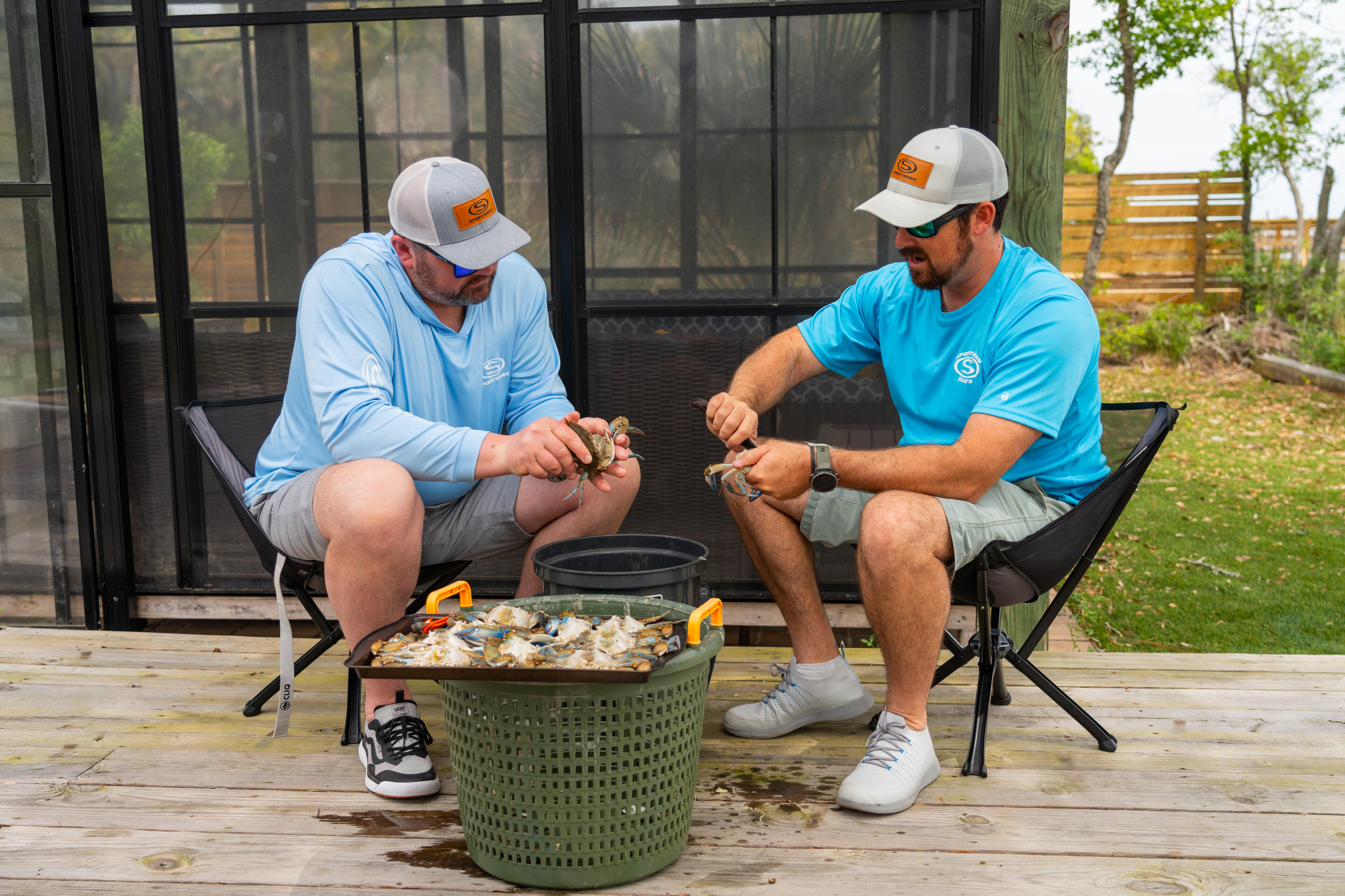 Two men in branded caps examine fresh crabs while seated on a wooden dock with a catch basket between them